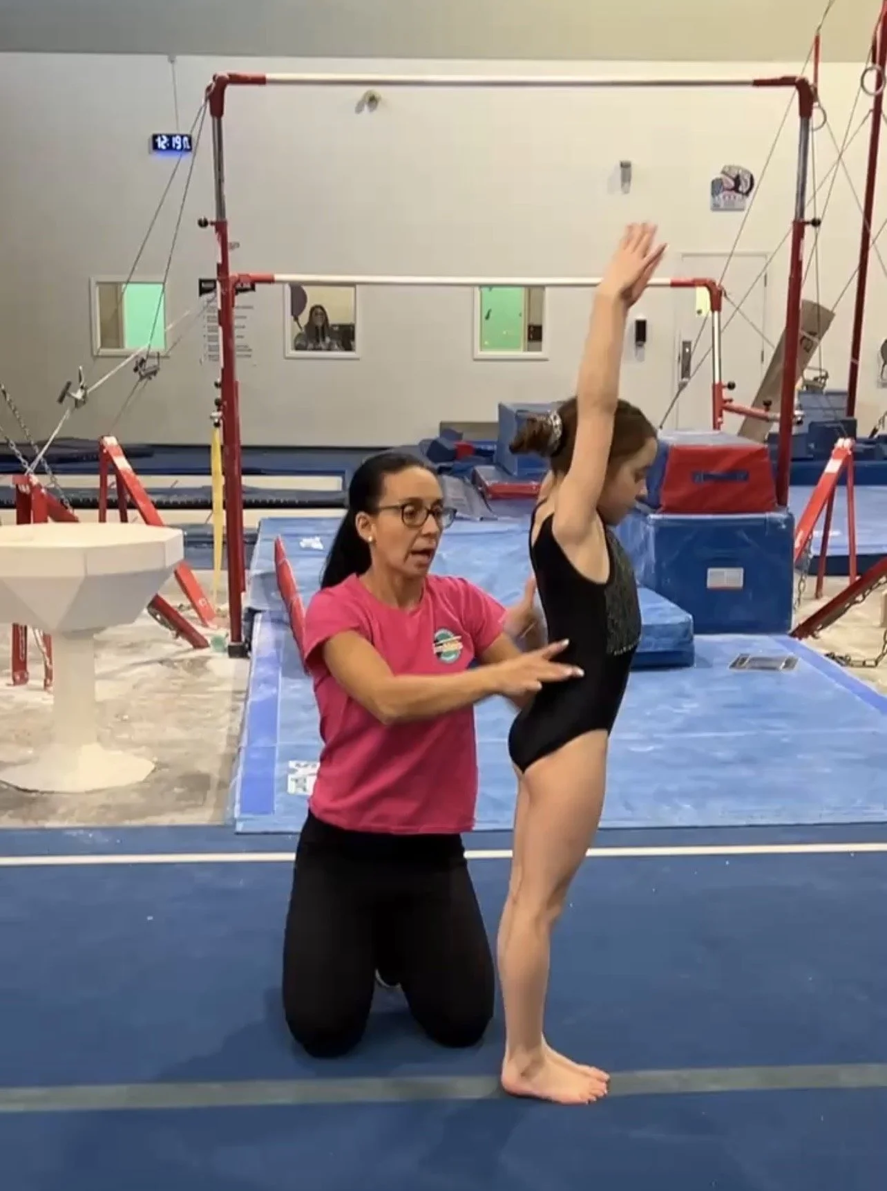 Coach in pink is ready to spot a female gymnast performing a skill.