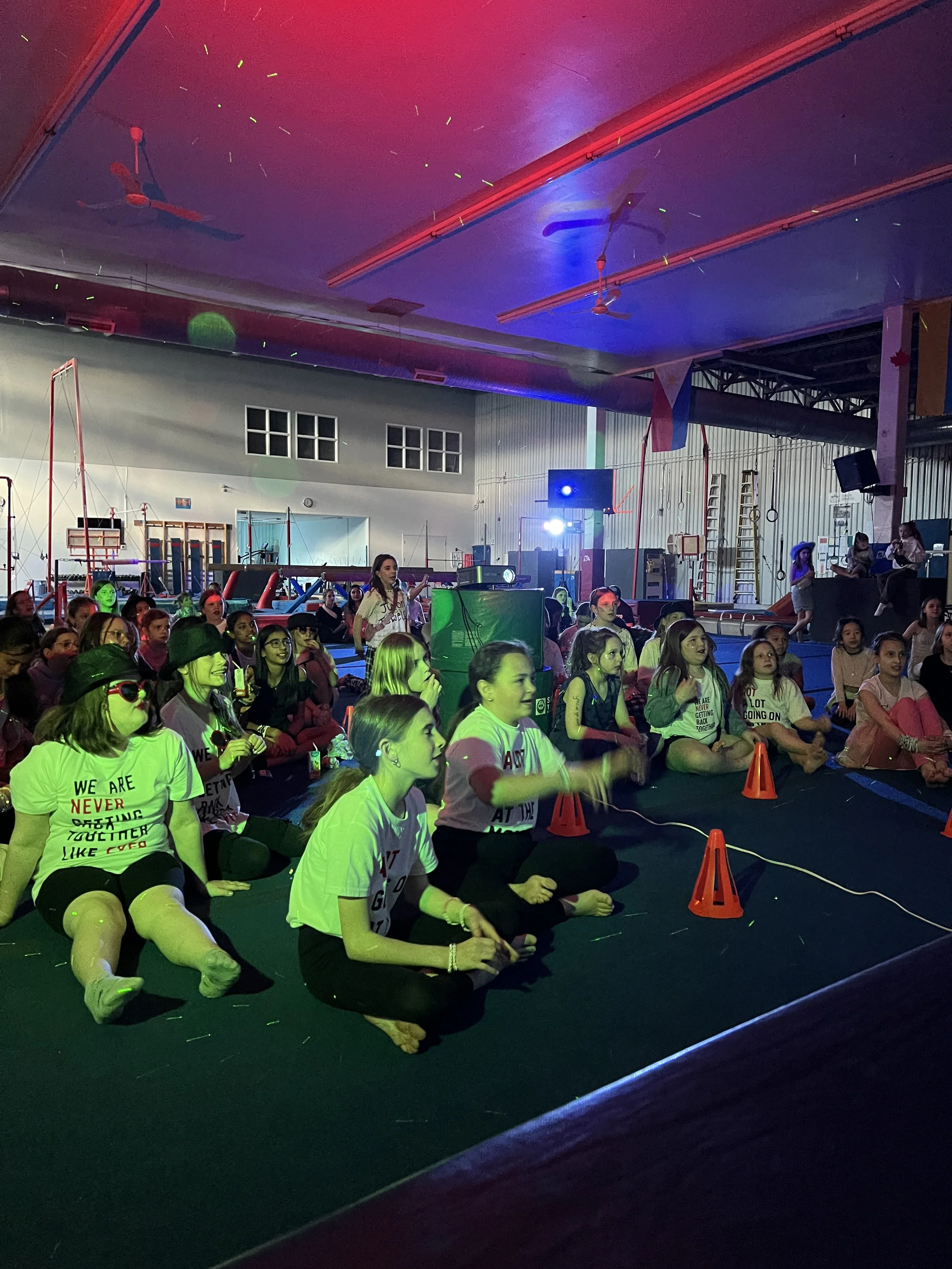 Many kids in the Tumblers Gymnastics Centre in the dark with glow in the dark lights all sitting down.