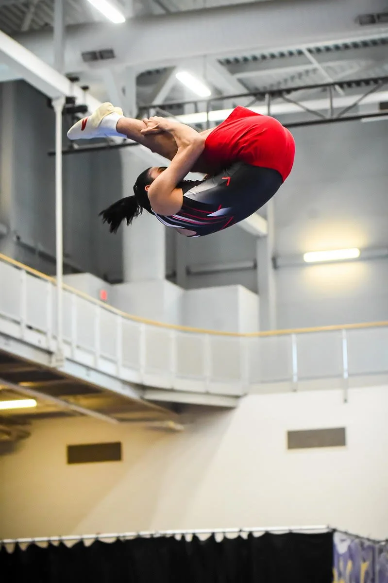 Man in black and red trampoline uniform does a flip in a pike position