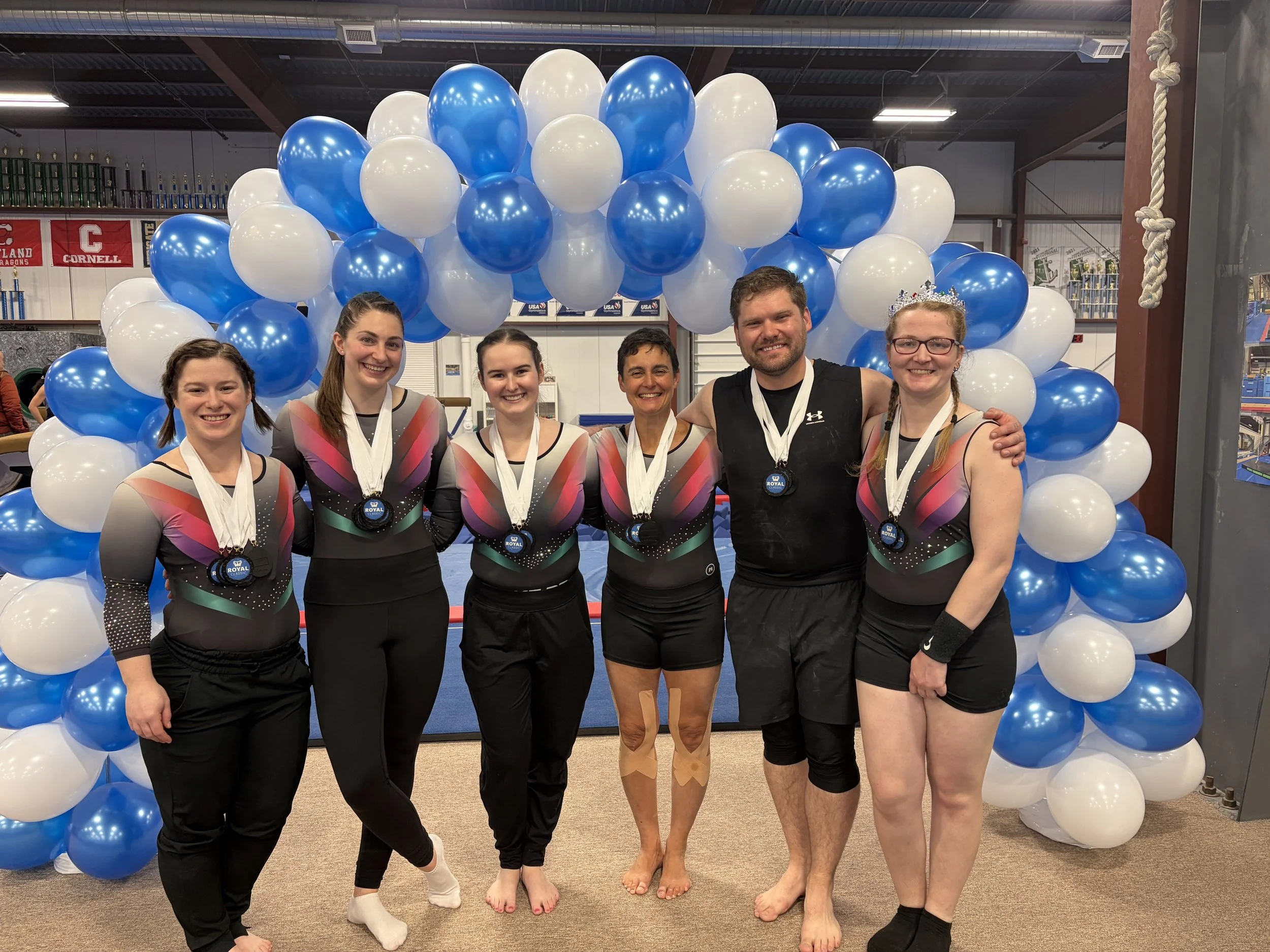 Group of adults standing arm in arm with medals around their necks. They are in gymnastics uniforms and just finished competing.