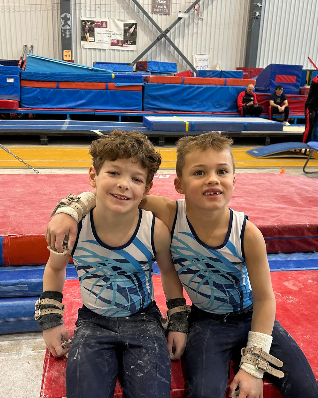 Two male gymnasts arm in arm smiling with gymnastics mats in the background