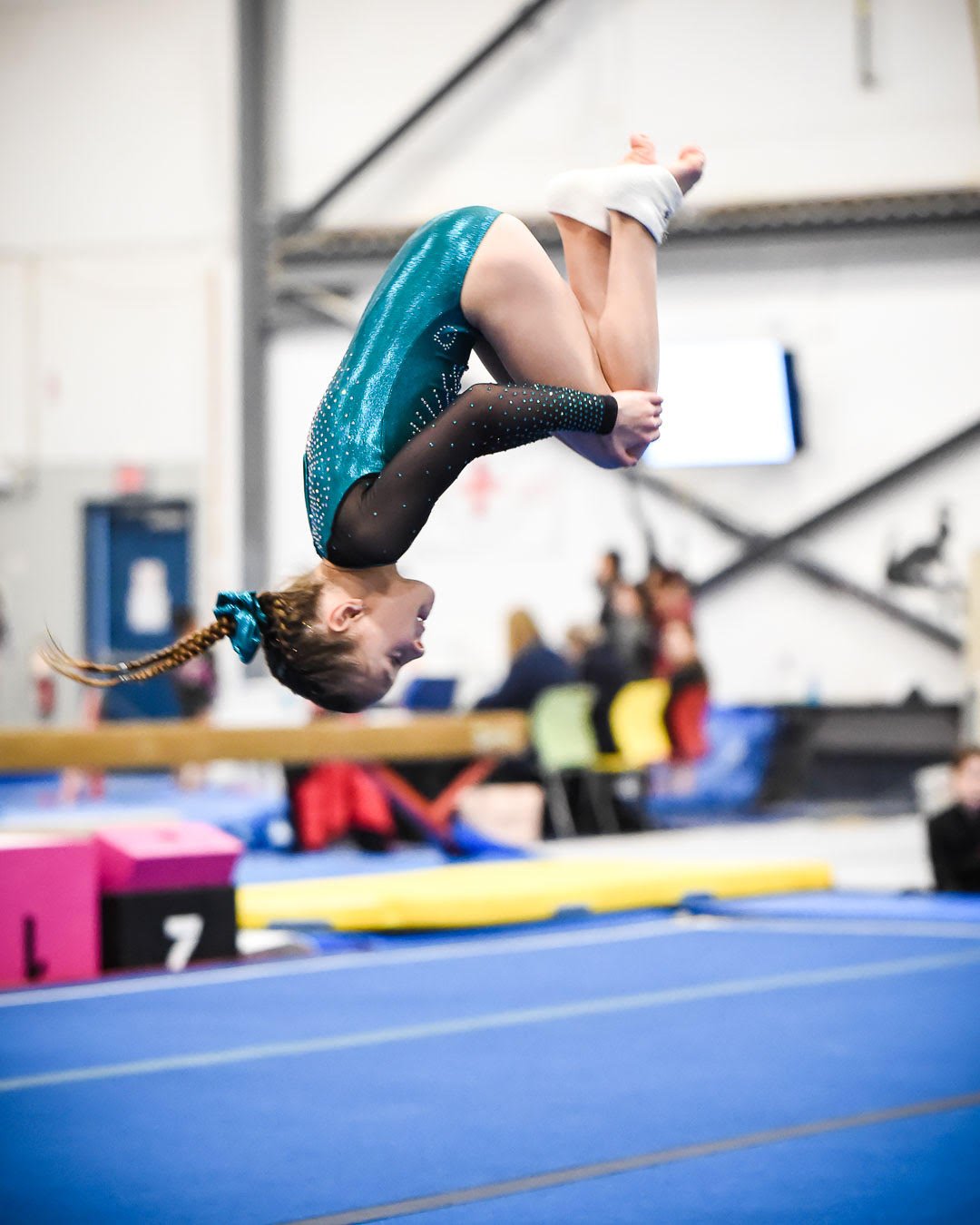 Girl in teal gymnastics leotard is performing a flip in a tuck position
