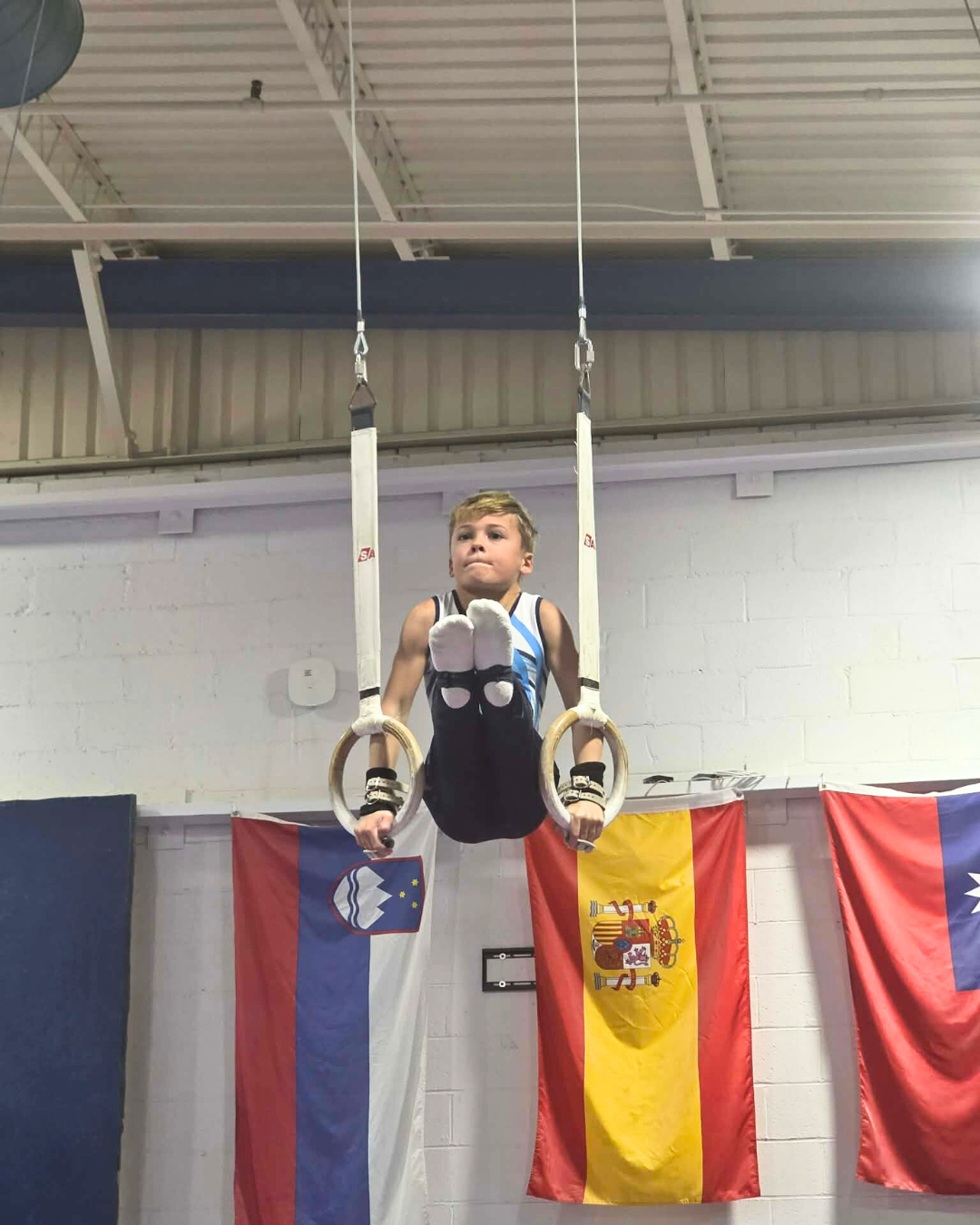 Young male gymnast on rings performing an L sit with country flags in the background