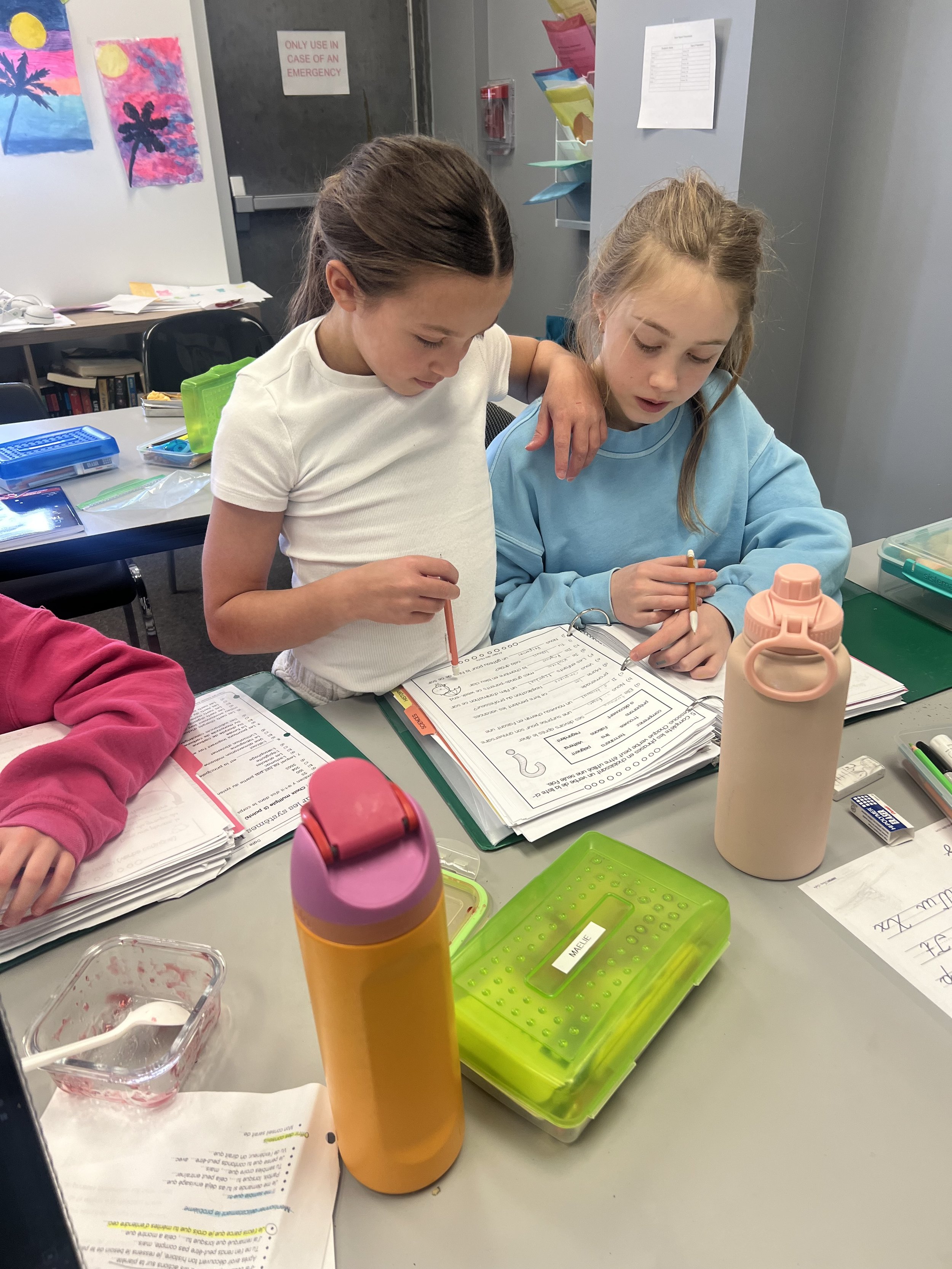 Two girls at a classroom desk looking at school notes. Image has two water bottles and a pencil case also on the desk.