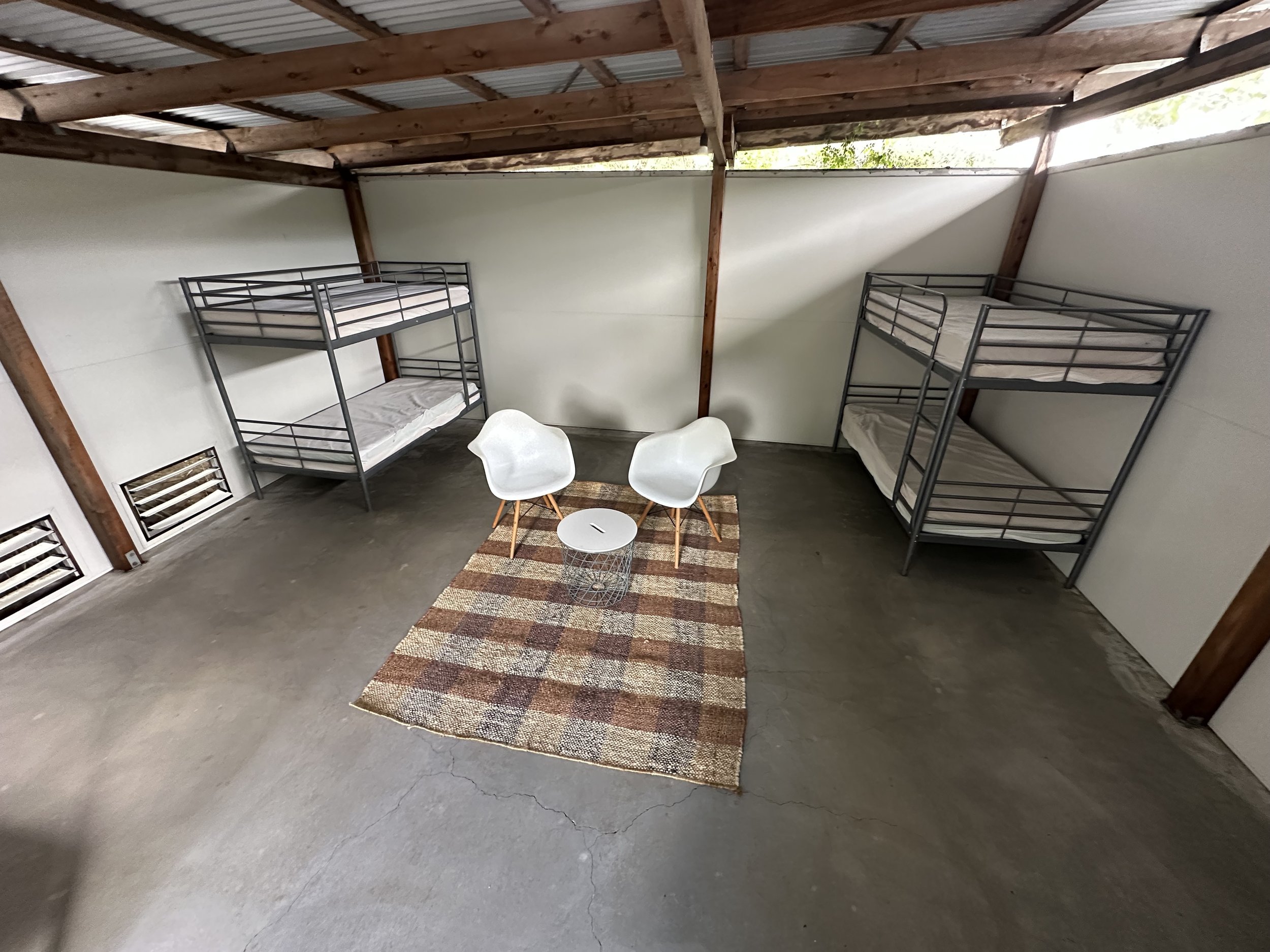 A room with wooden beams and a metal roof, containing metal bunk beds with mattresses, a small area rug, two white chairs, and vents on the wall. The Regenerative Training Centre, Woodford, Qld.