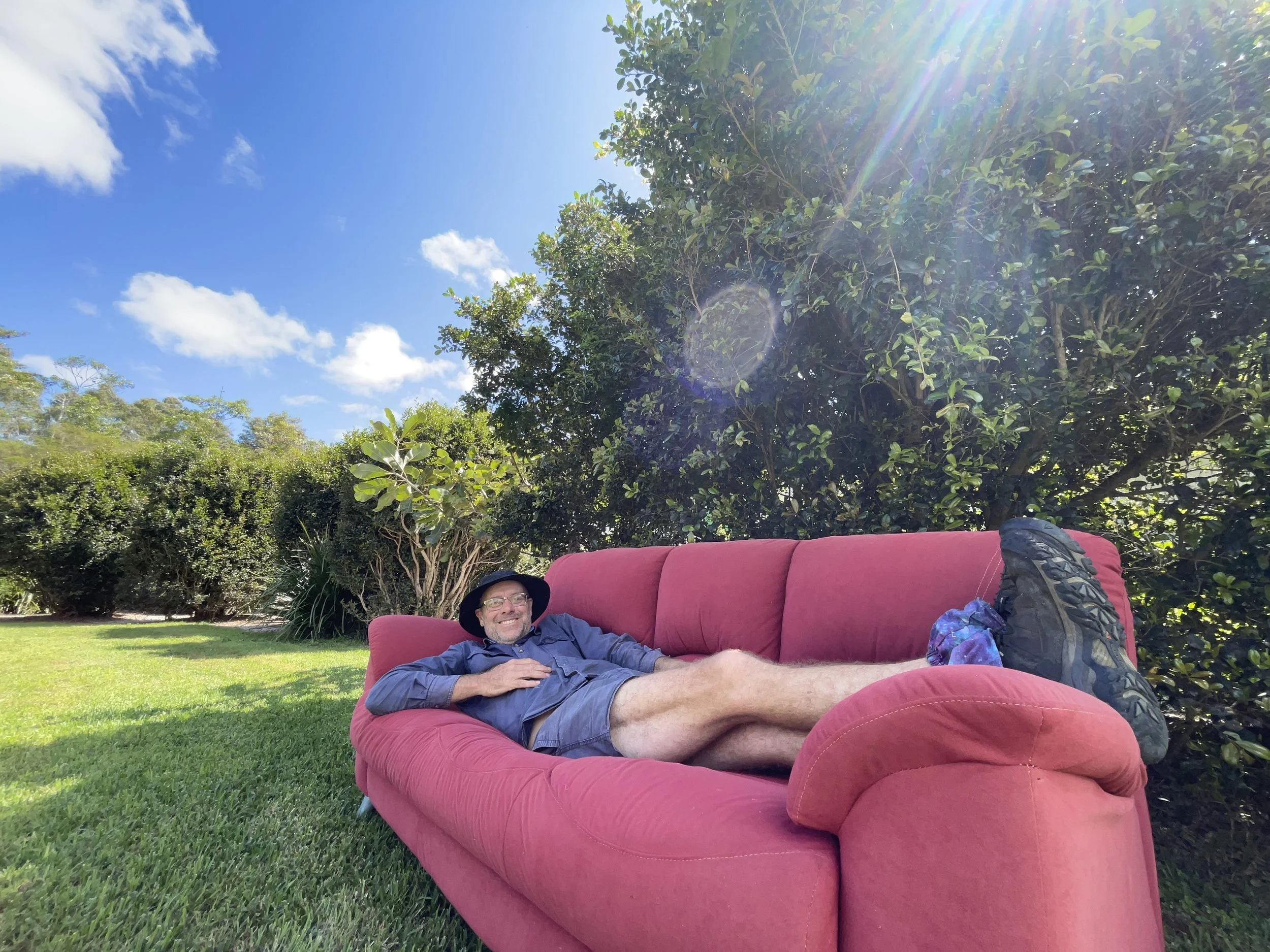 A man lying on a red outdoor sofa on grass, wearing glasses, a hat and a long-sleeved shirt, smiling at the camera. The background features green trees and a bright blue sky with some clouds and sunlight. The Regenerative Training Centre, Woodford, Q