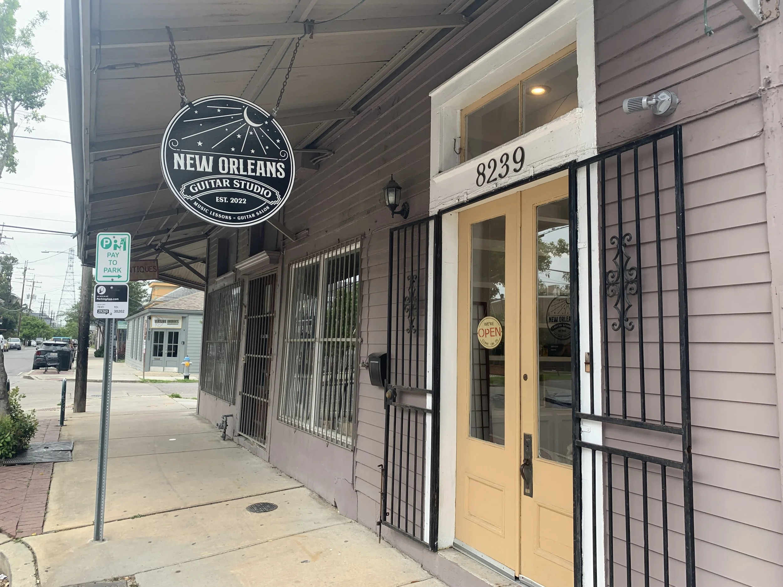 Exterior view of a building with a pizza sign that reads "New Orleans Guitar Studio, Est. 2022, Music Lessons - Guitar Salon," along with the street number 8239 and a glass door with a small sign that says "We're Open." The building has barred windows, and there is a parking sign on the sidewalk indicating paid parking.