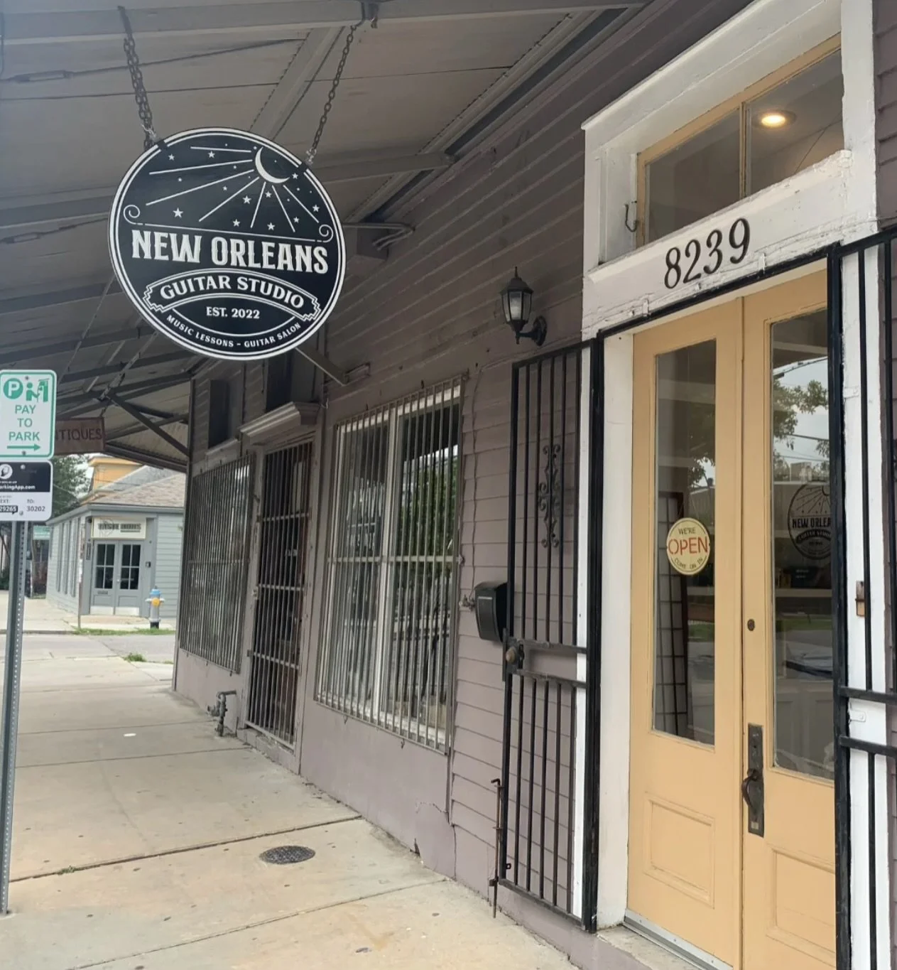 Exterior of a music lessons and guitar salon studio named 'New Orleans Guitar Studio' at 8239, with a sign indicating the business was established in 2022. The storefront has a yellow door with a round 'We're Open' sign and is secured by black metal bars on the windows and door.