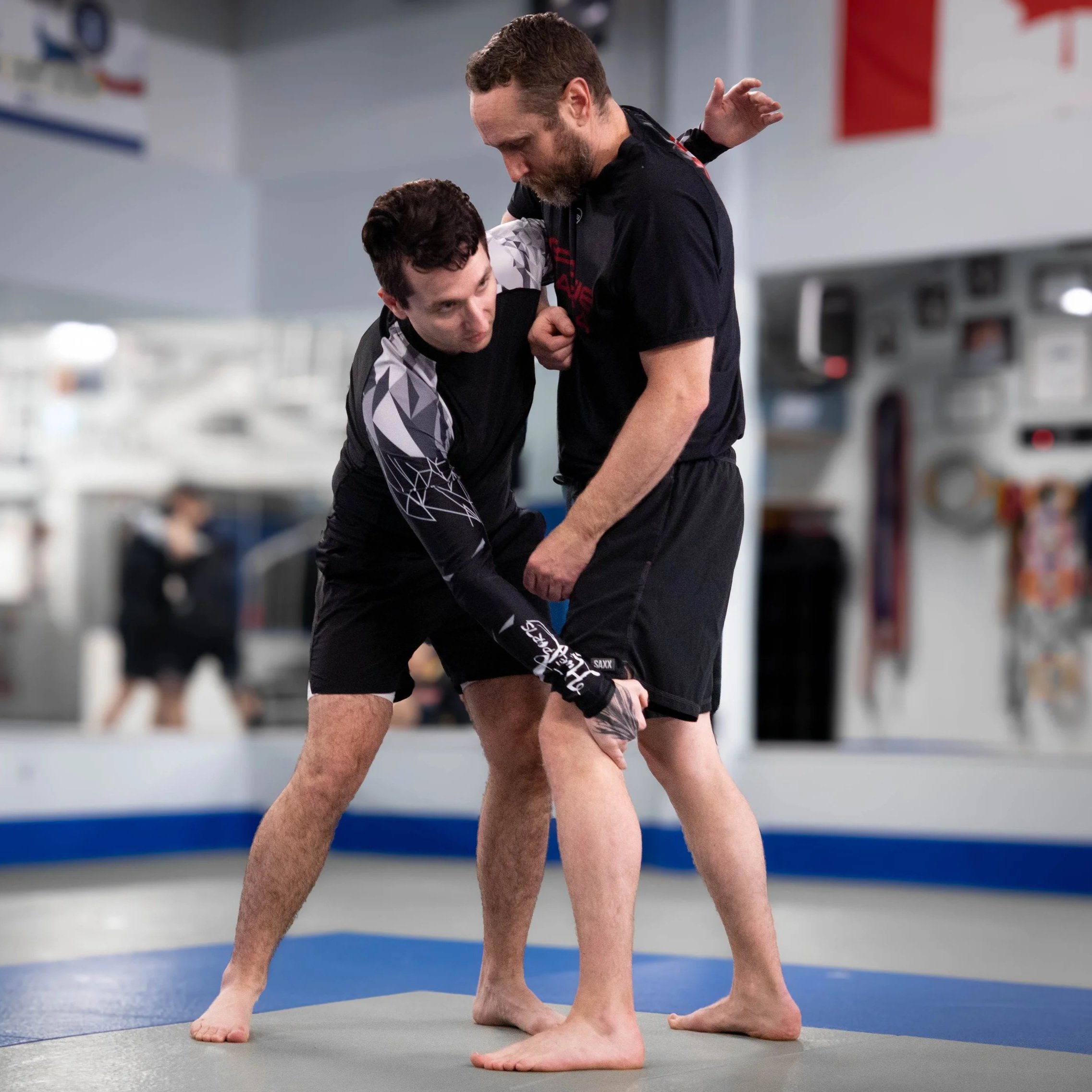 Two people practicing Brazilian Jiu-Jitsu on a mat, with one person standing and the other on the ground wearing gis.
