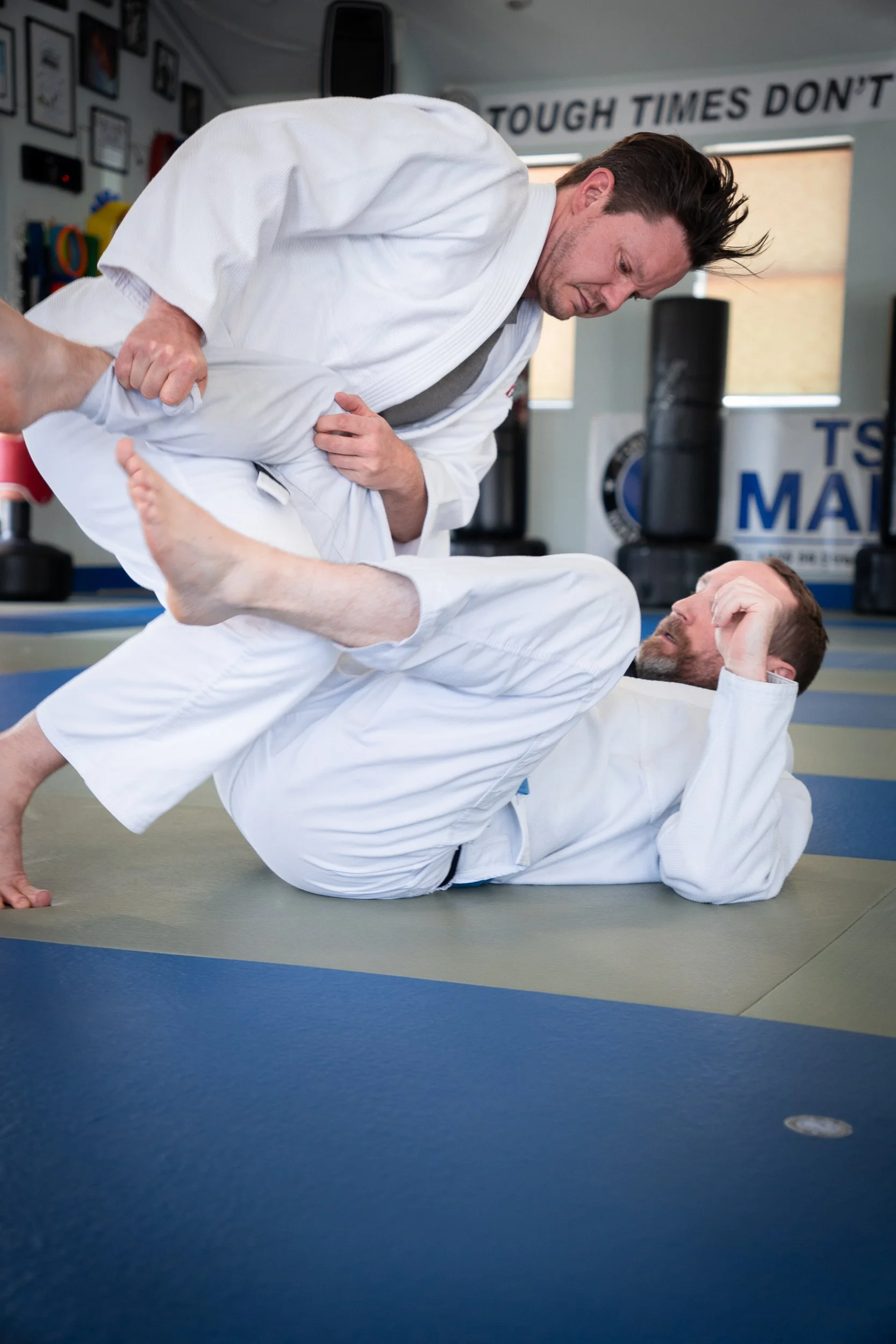 Two individuals practicing Brazilian jiu-jitsu on a padded floor, one wearing a blue gi and the other a green gi. Background shows onlookers seated.