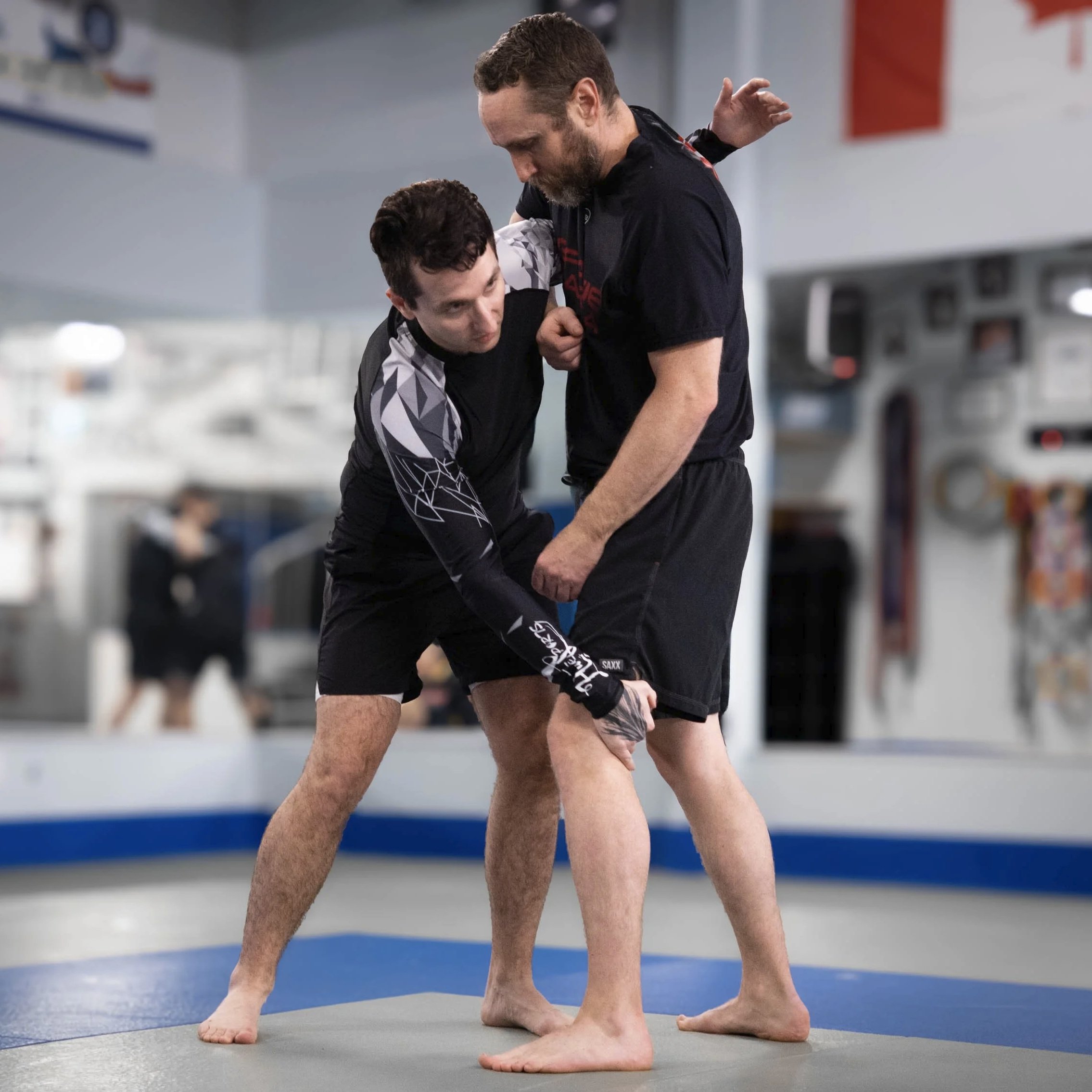Two people practicing Brazilian Jiu-Jitsu on a mat, with one person standing and the other on the ground wearing gis.