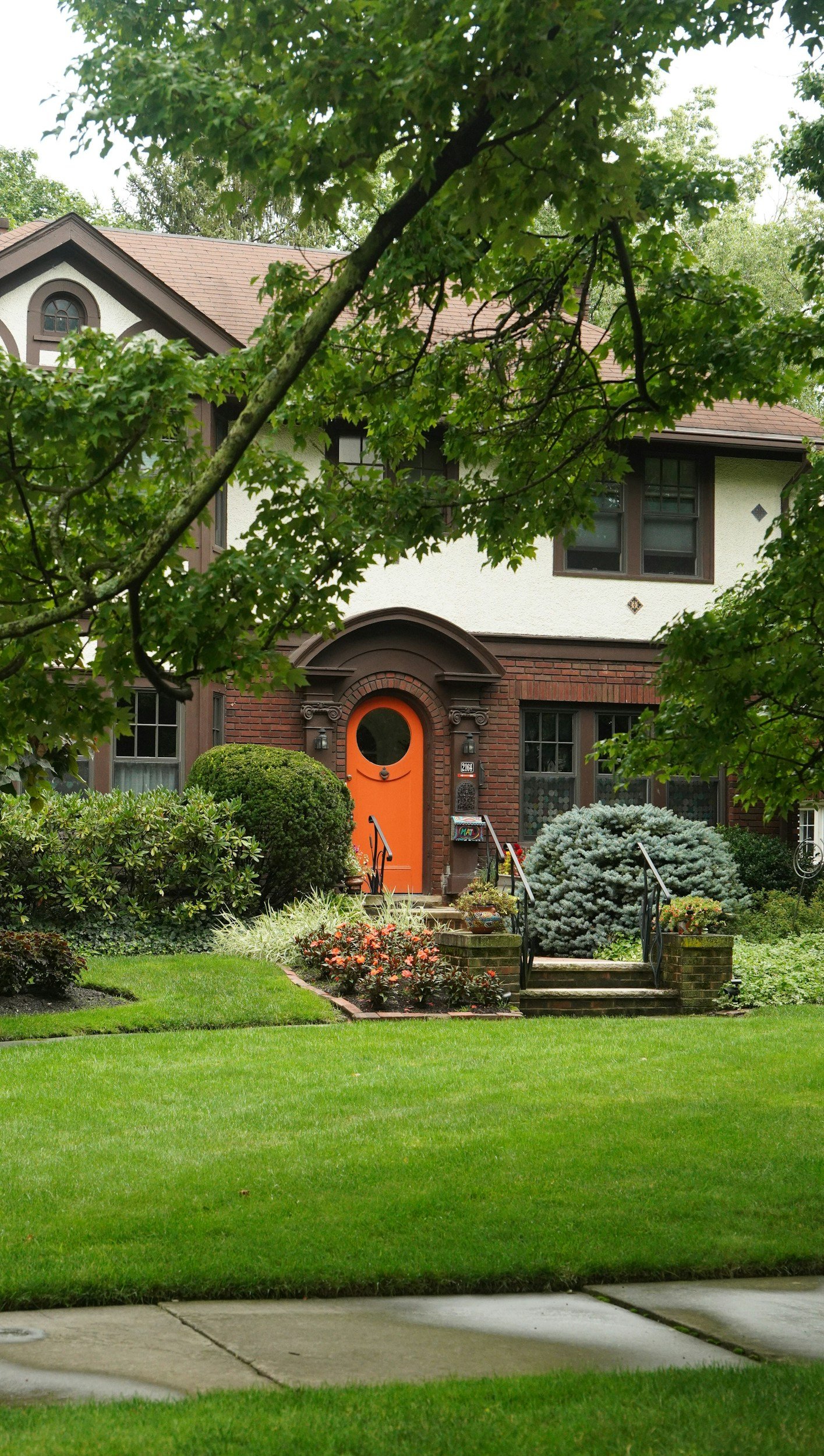 Front view of a two-story house with an orange door, surrounded by a landscaped garden and green lawn, partially obscured by tree branches.