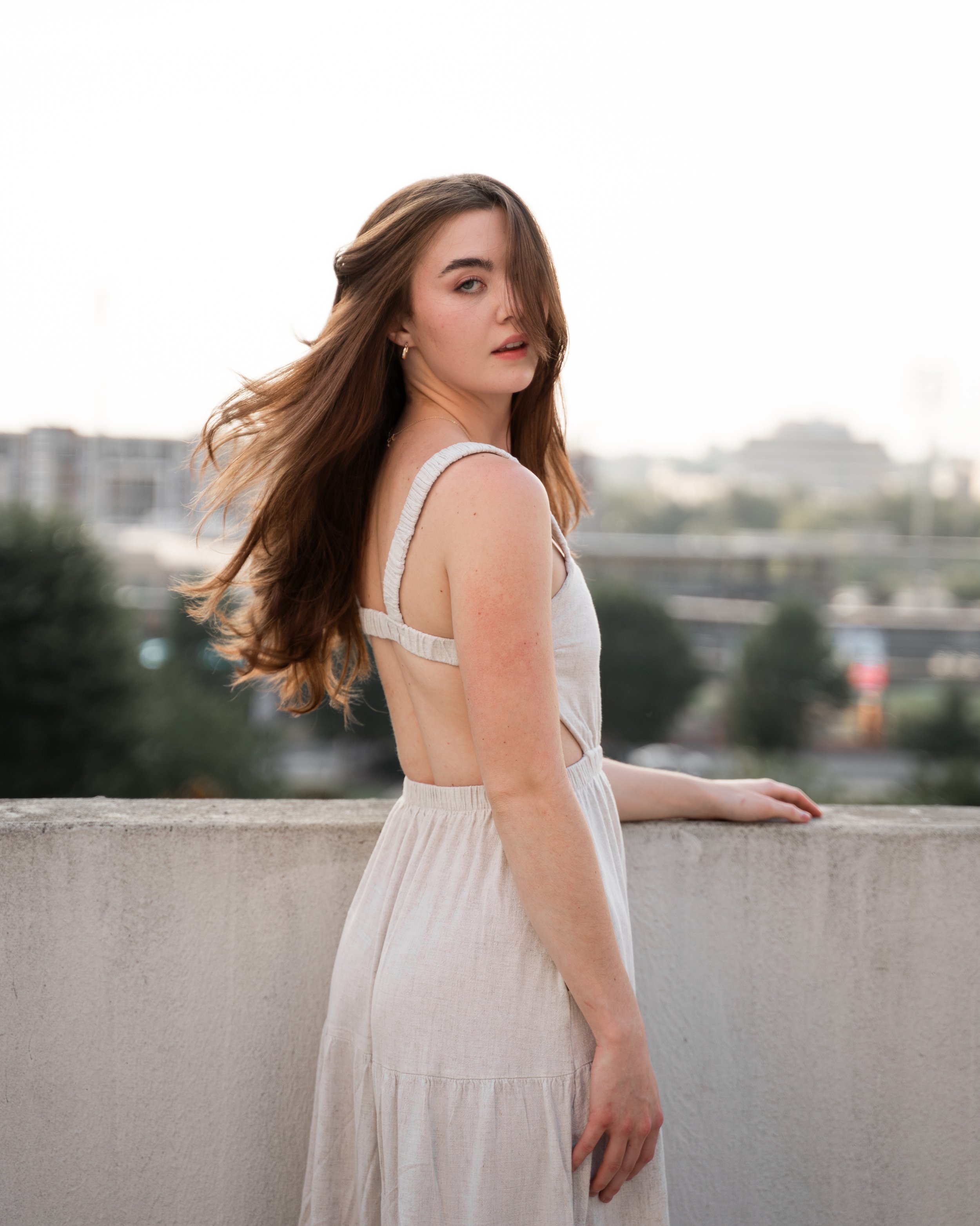 Woman in a light dress standing on a rooftop with hair flowing and a blurred cityscape in the background.