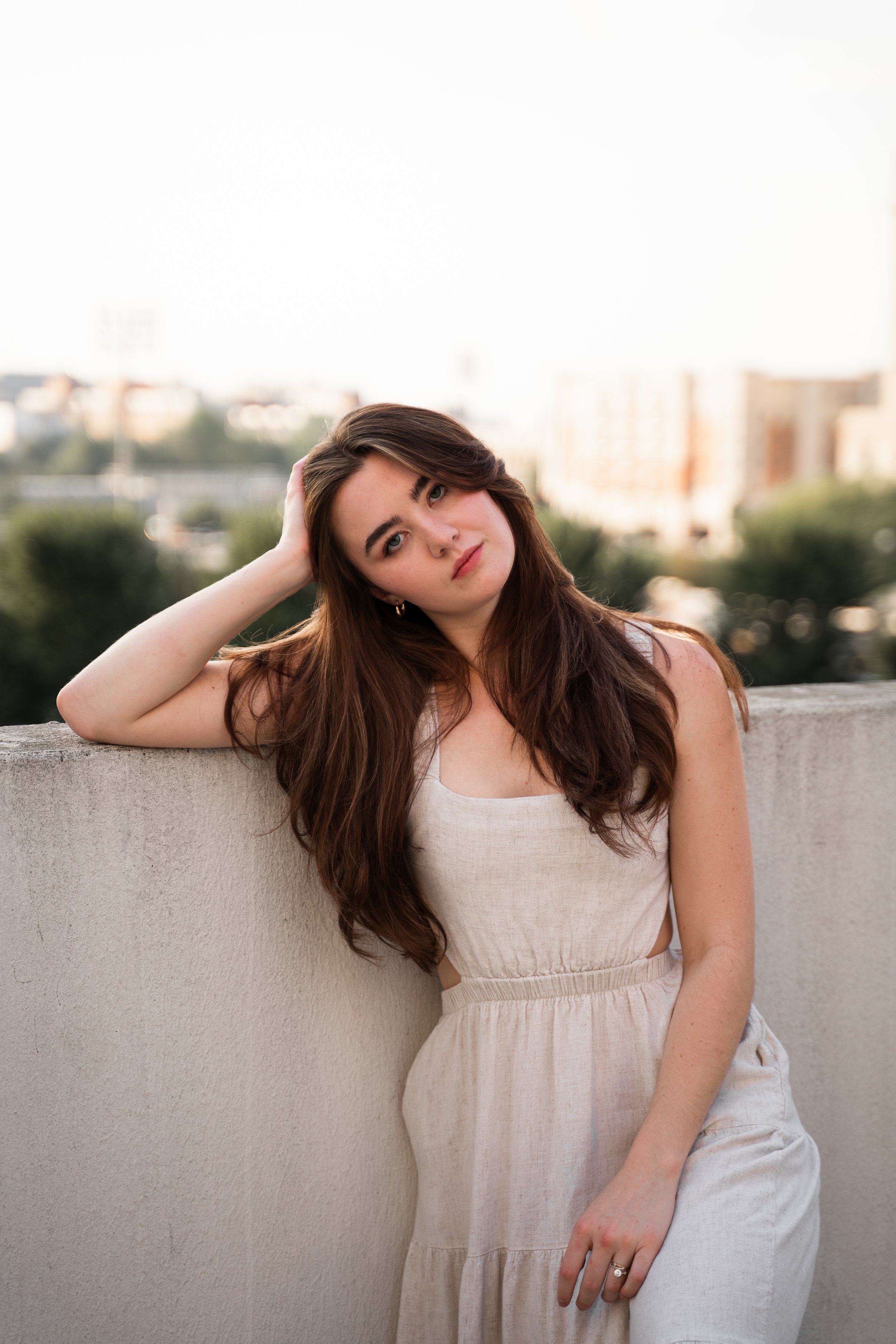 Woman in a beige dress leaning on a wall outdoors with a cityscape in the background.