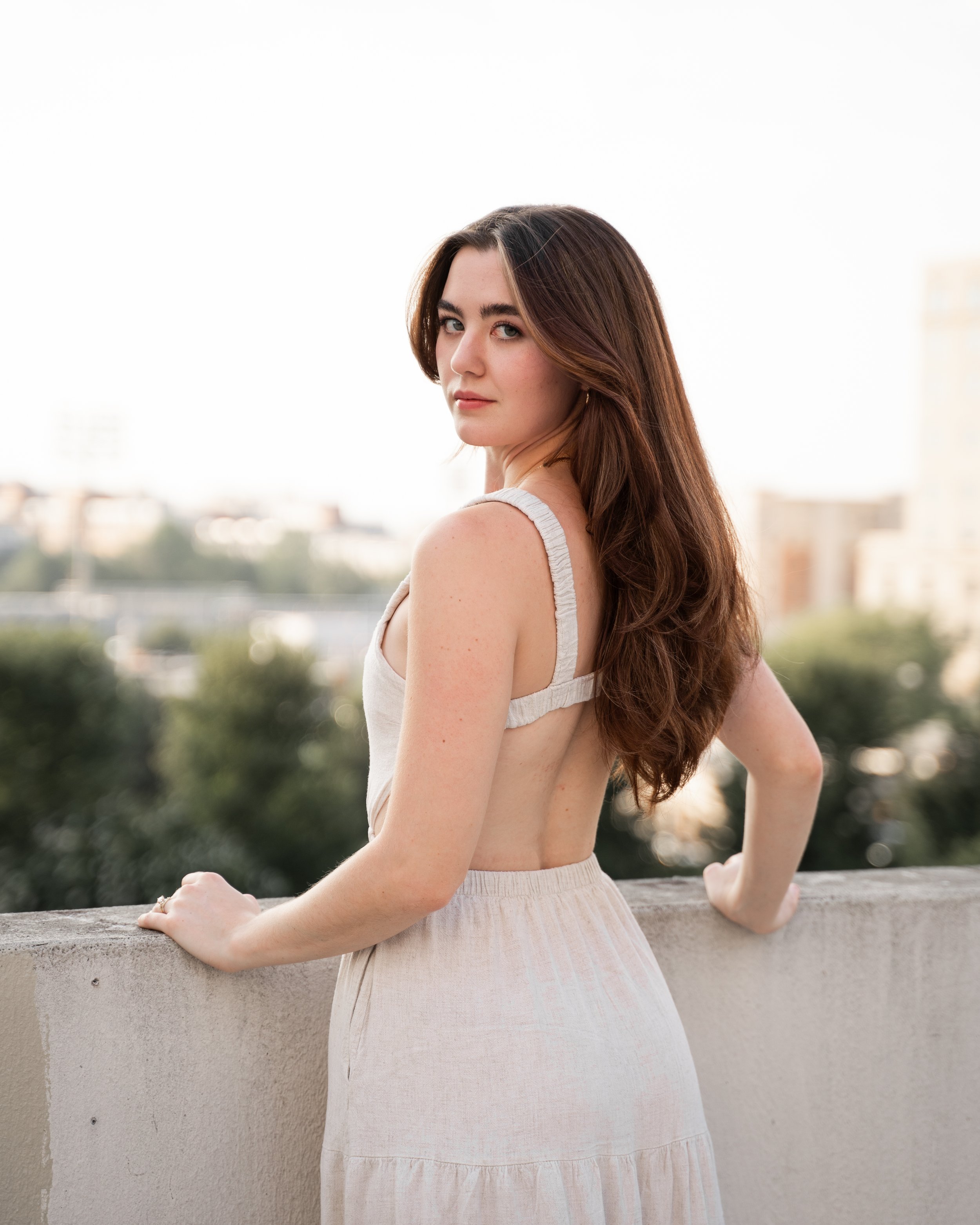 A woman with long brown hair wearing a sleeveless, backless dress stands by a concrete wall outdoors, looking over her shoulder at the camera.