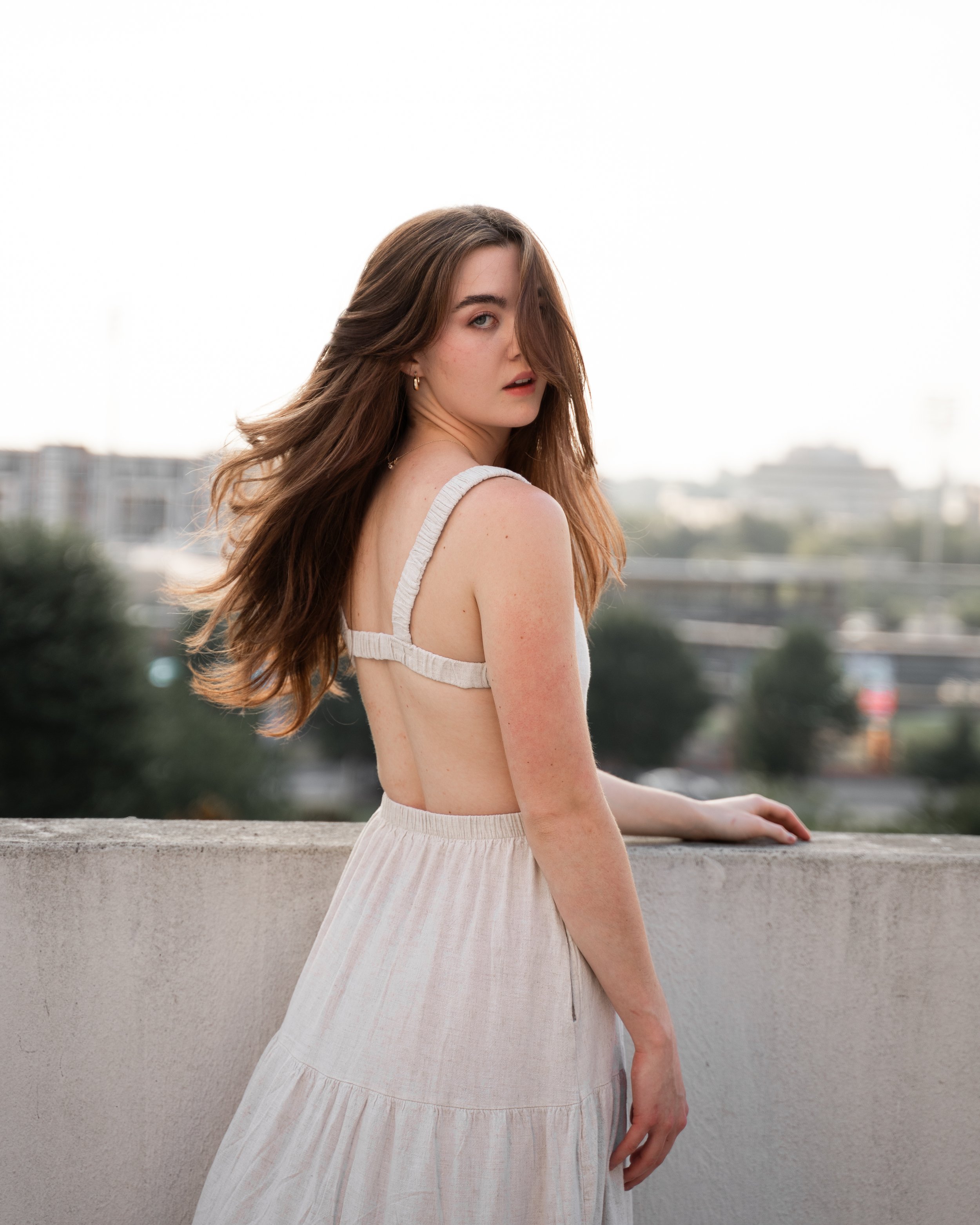 A woman in a sleeveless white dress with open back standing by a concrete wall outdoors, with the city skyline in the background.