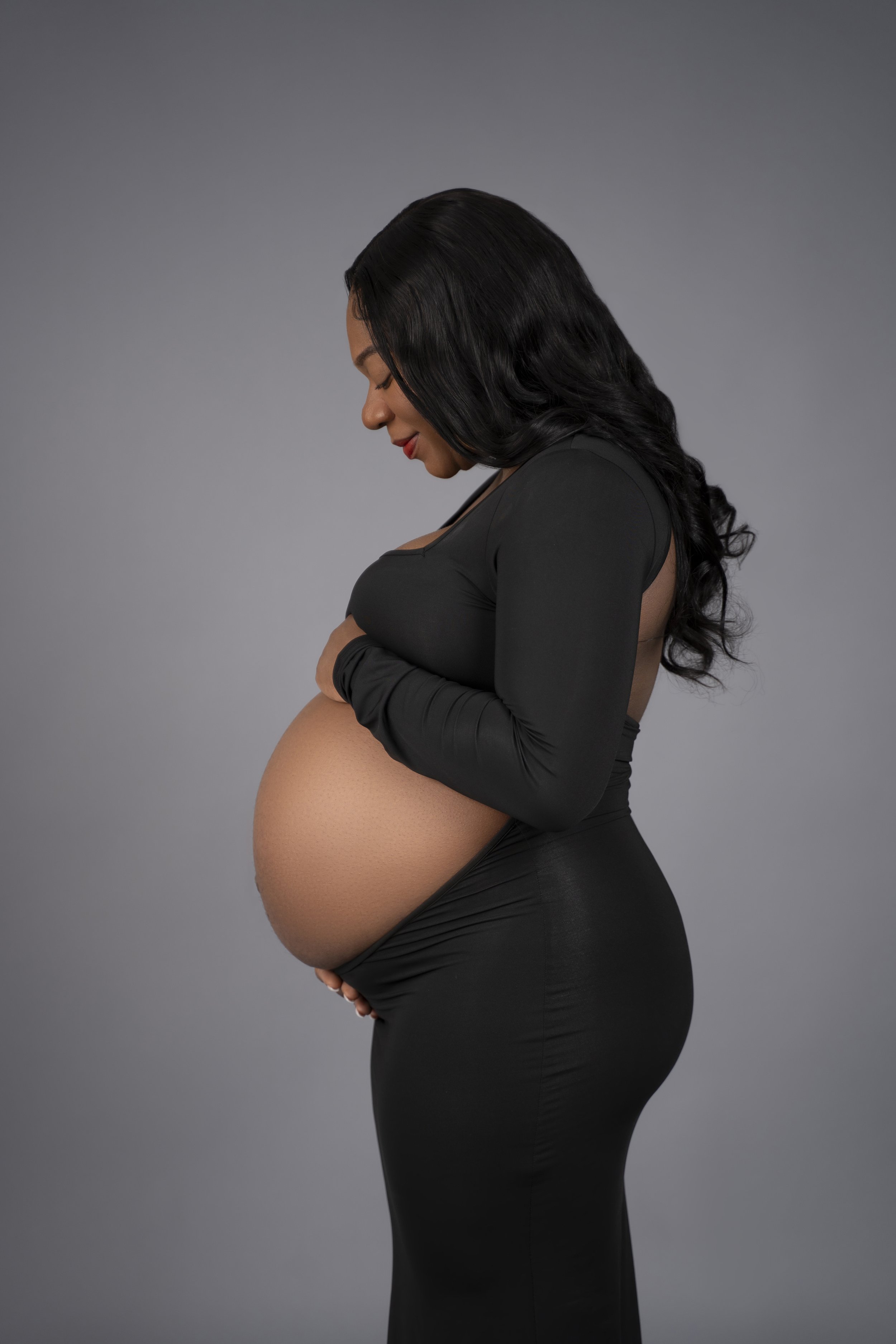 Maternity studio photoshoot in a black dress holding her belly, profile view.
