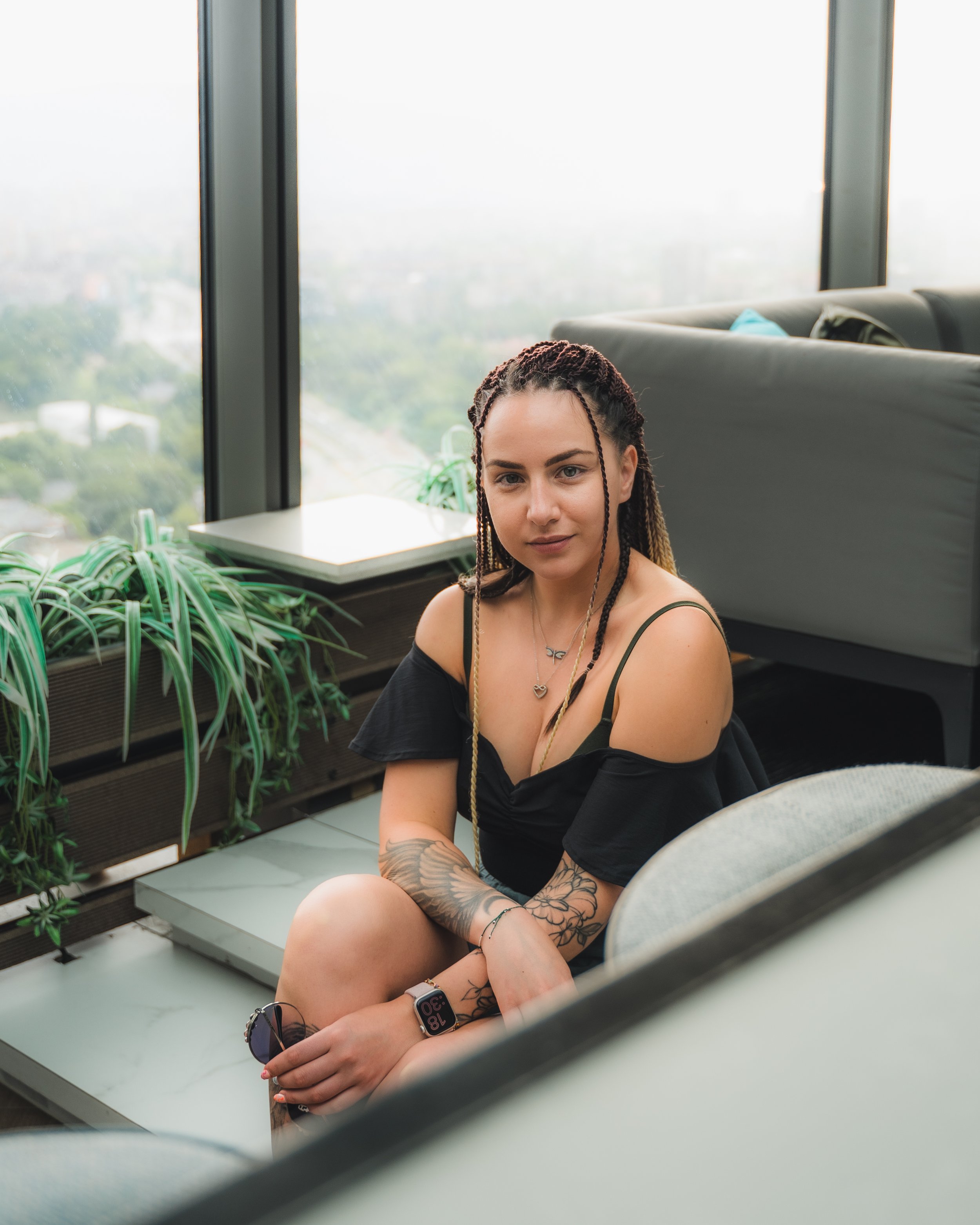 Woman with braided hair sitting indoors, wearing a black top and smartwatch, next to large windows with a cityscape view.