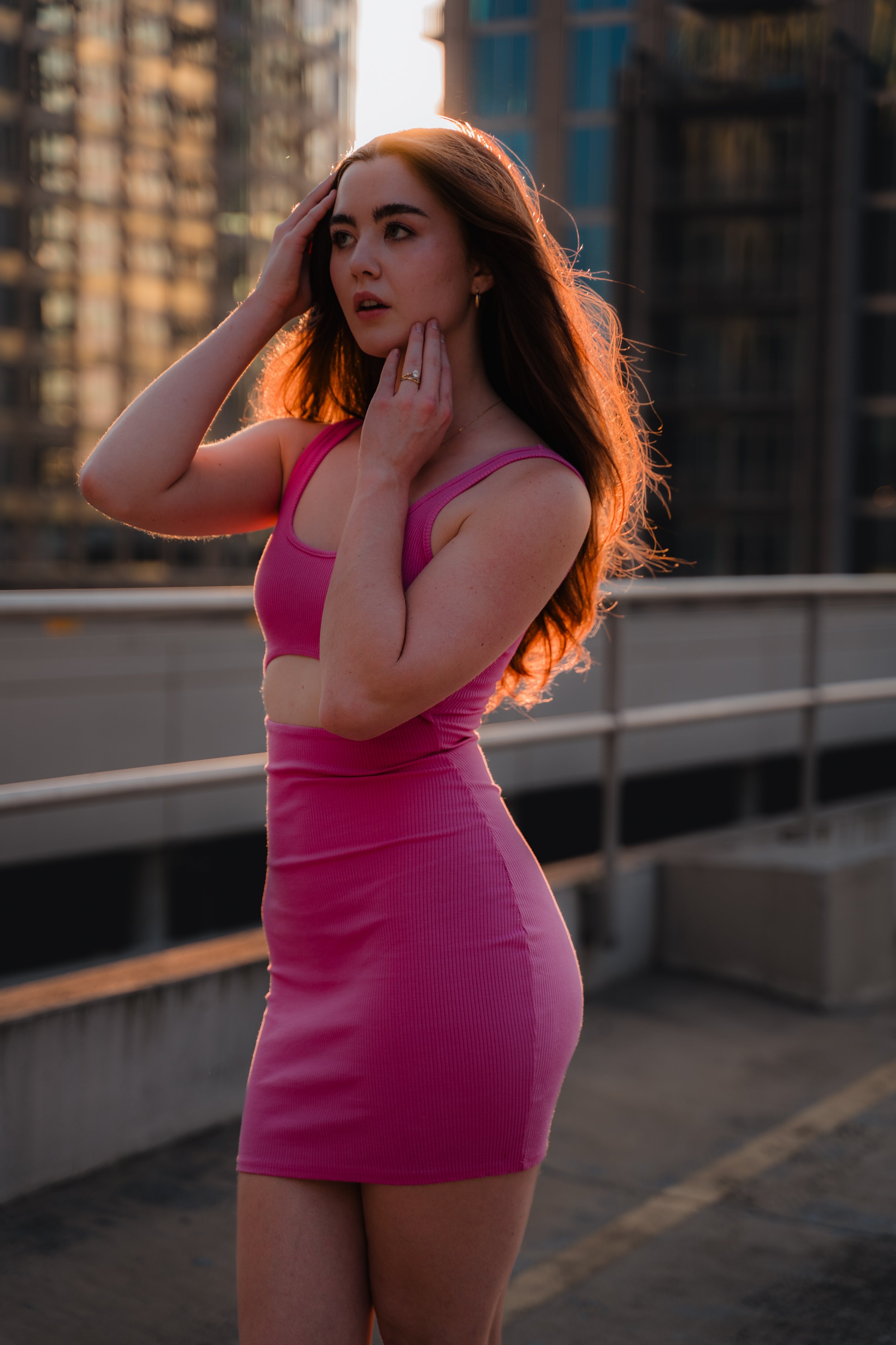 Woman in a pink dress standing on a rooftop with city buildings in the background at sunset.
