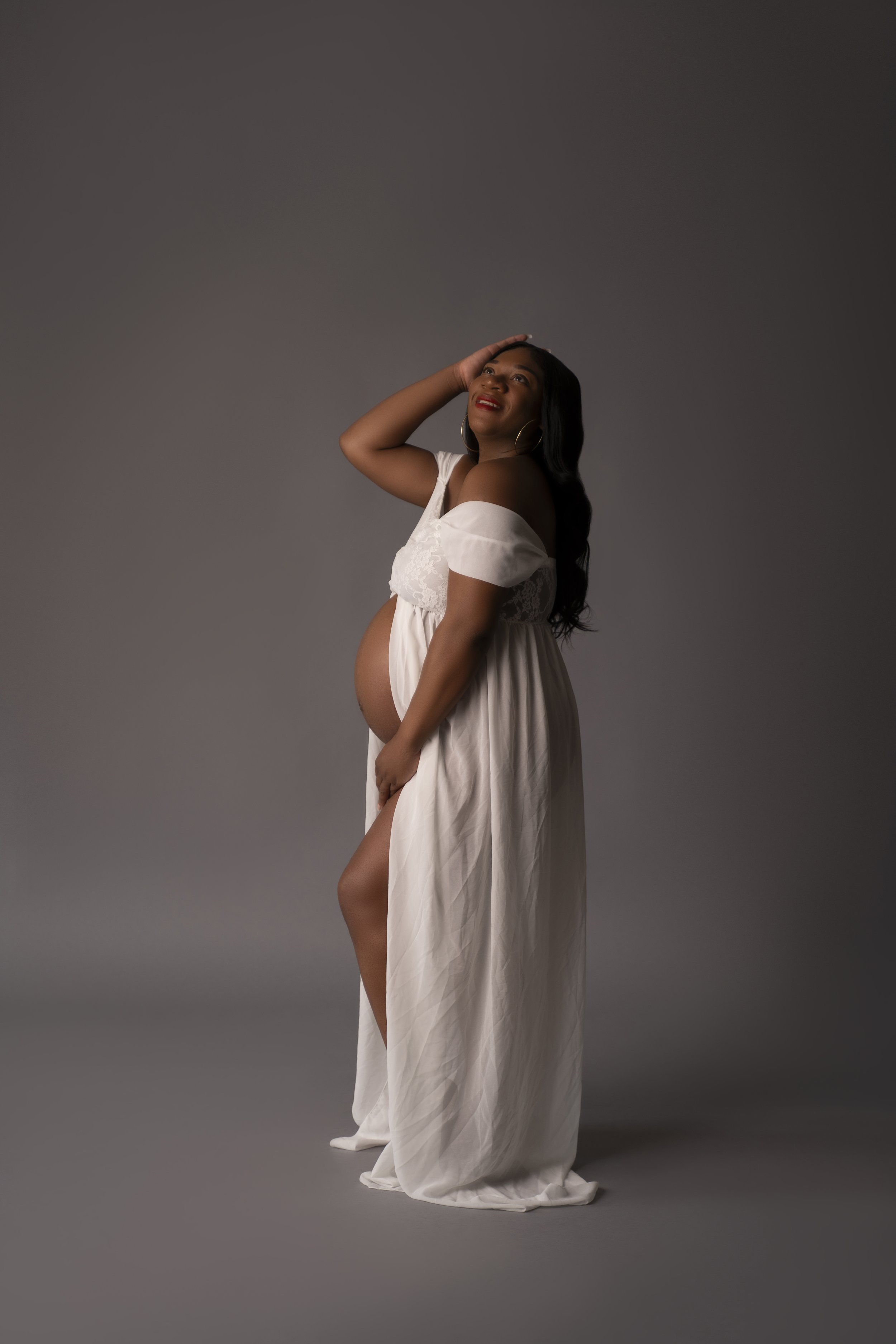 Maternity portrait of a woman posing in white dress against gray background.