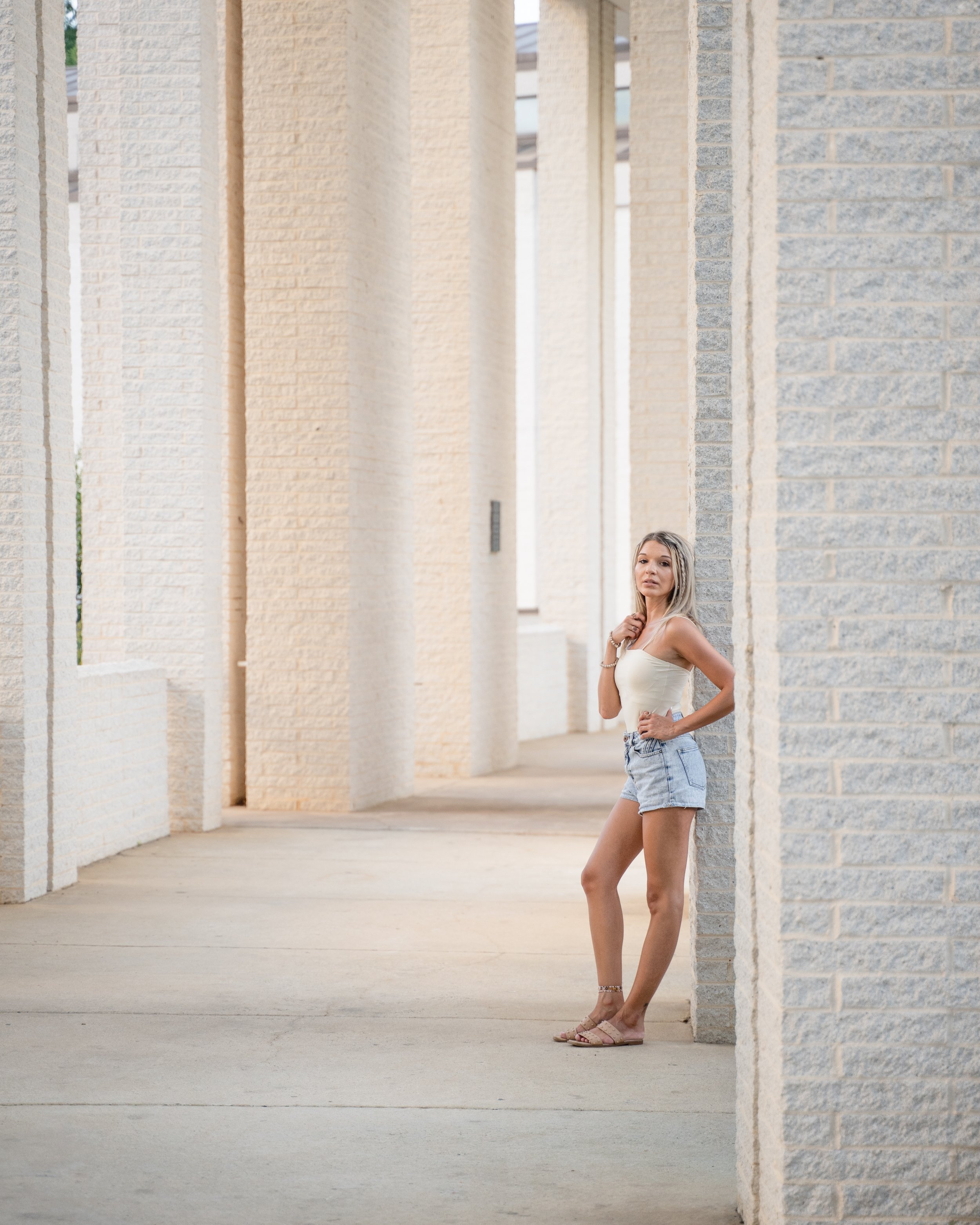 A woman in a white top and denim shorts stands in a corridor with textured white brick walls and columns, posing for a photo.
