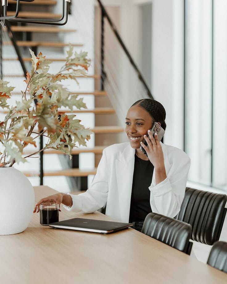 Businesswoman in white blazer talking on phone, seated at a wooden table with a laptop, drink, and plant, stairway in background.