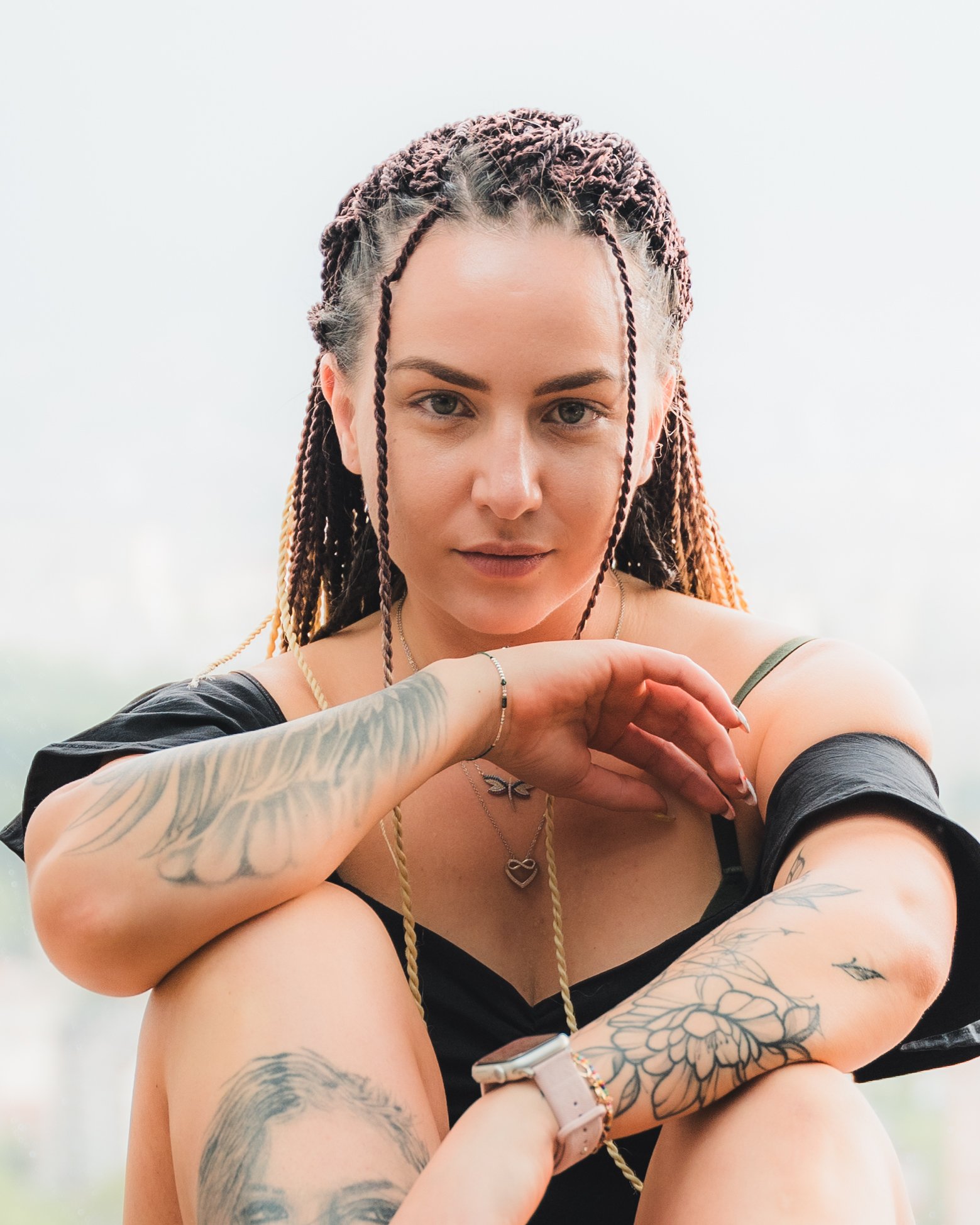 Woman with braided hair and tattoos sitting with a relaxed posture, wearing a black off-shoulder top.