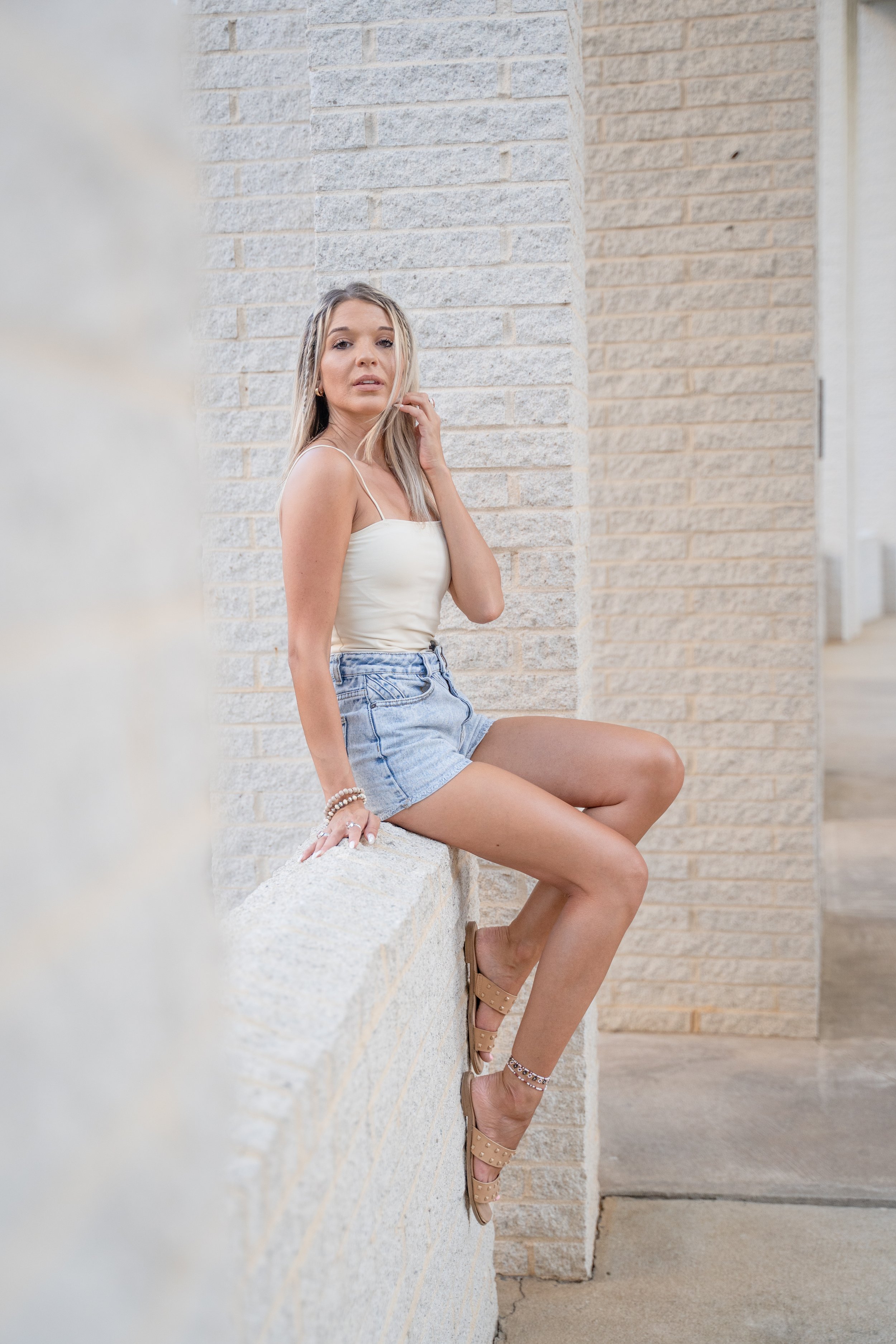 Woman sitting on a white brick ledge wearing a white tank top, denim shorts, and sandals, with a neutral expression, against a brick wall backdrop.