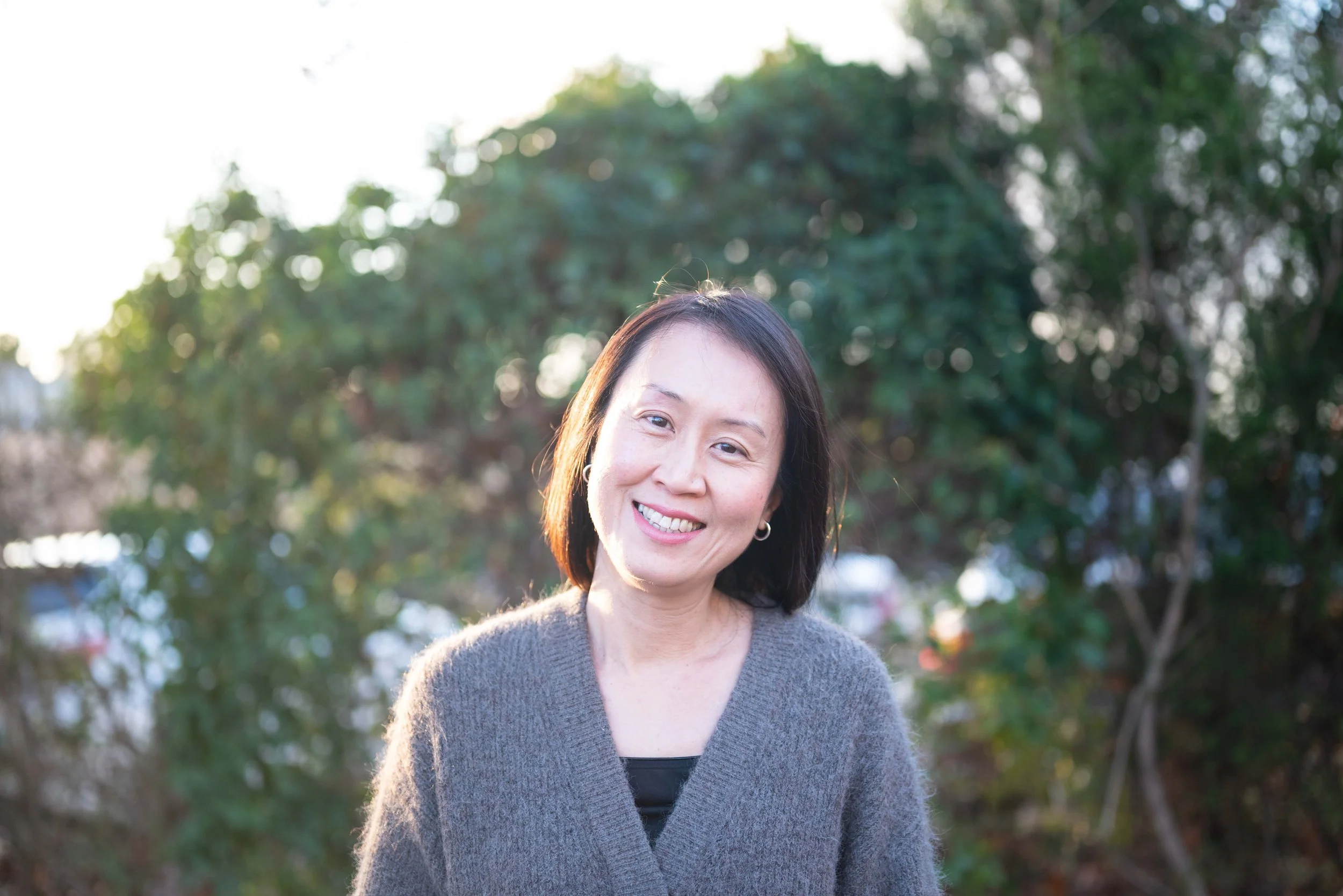 A woman with dark hair smiling outdoors with greenery and sunlight in the background.