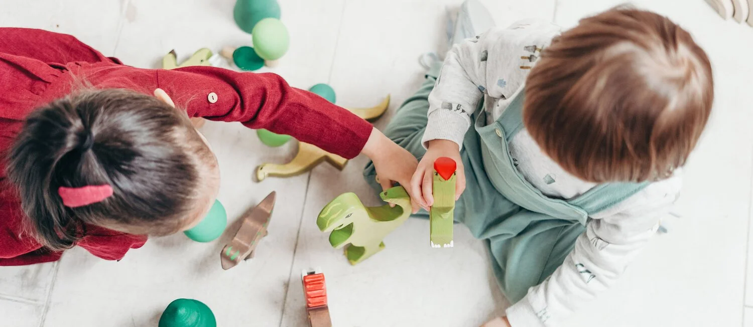 Two children playing with wooden toys on a light-colored floor, surrounded by small green plastic balls.