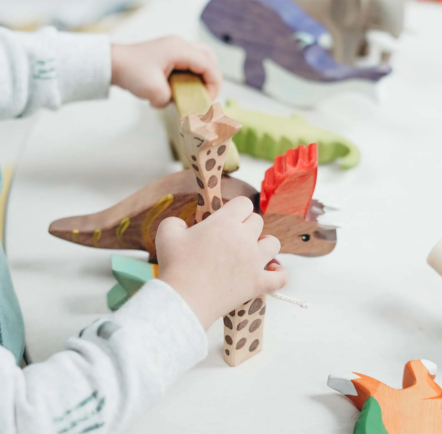 Child's hands playing with hand-painted wooden animal toys, including a giraffe and a dinosaur, on a white surface.