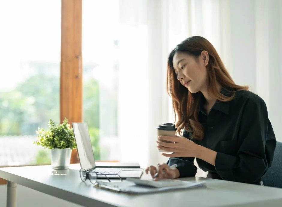 A woman with long brown hair sitting at a white desk in a bright room, holding a to-go coffee cup, with a laptop, a plant, and glasses on the desk.