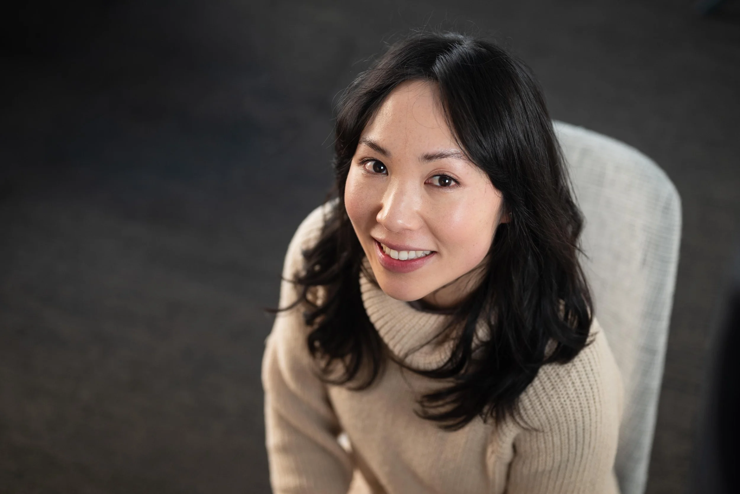 A woman with black hair smiling and looking up at the camera, sitting on a light-colored checked chair against a dark background.