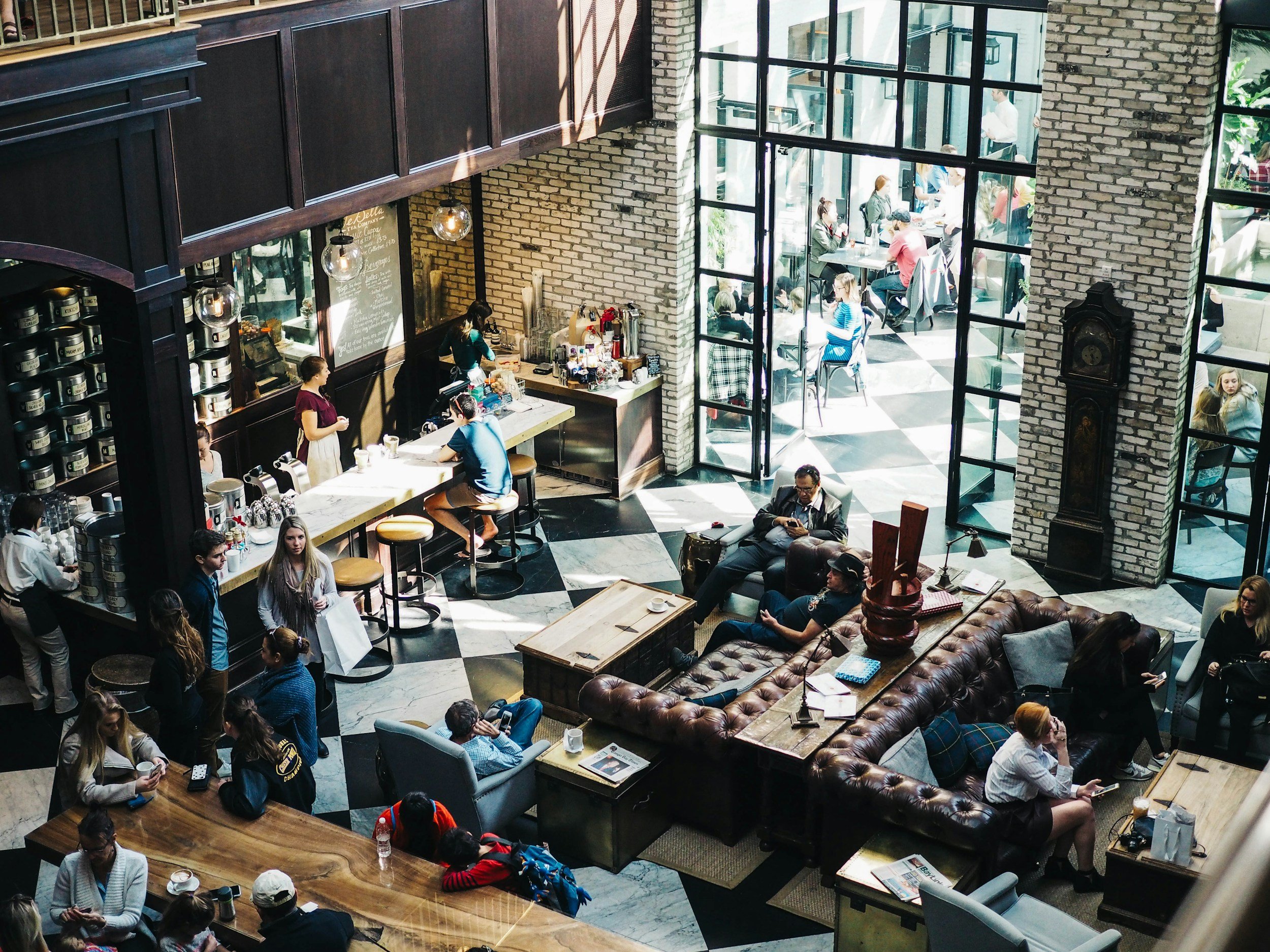 People sitting and standing in a modern, lively coffee shop with large windows, brick walls, and black-and-white checkered floors.