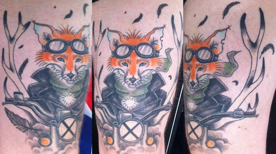 Tattoo of a fox wearing goggles and a leather jacket, riding a motorcycle with antlers in the background.