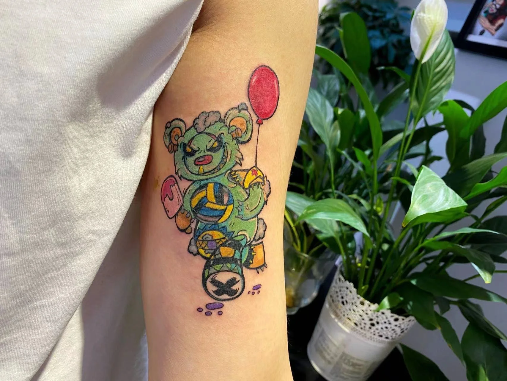 Colorful cartoon bear tattoo holding a red balloon on a person's arm with plants in the background.