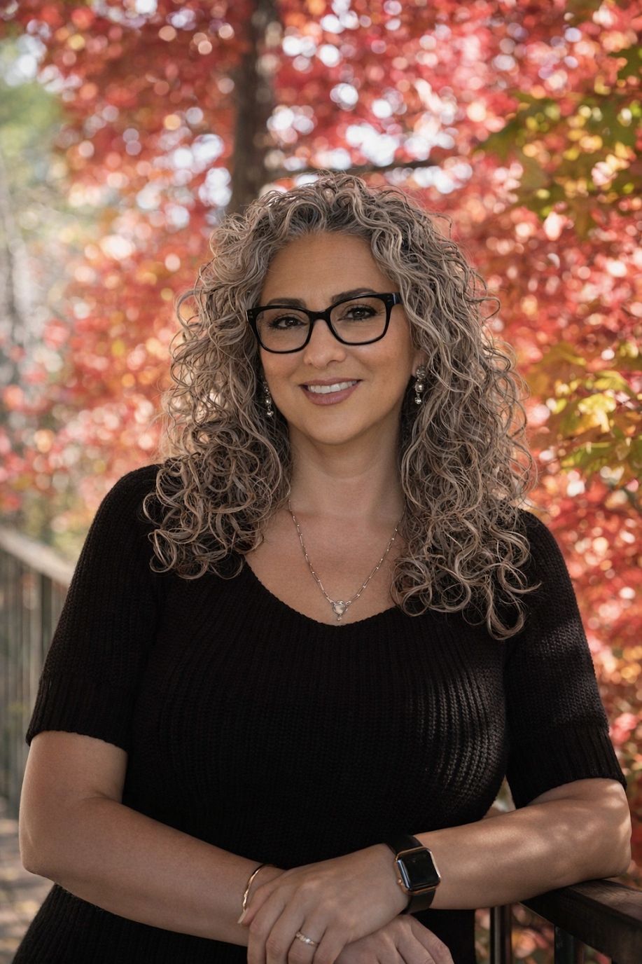 A woman with curly blonde hair, wearing black-rimmed glasses, smiling in front of greenery