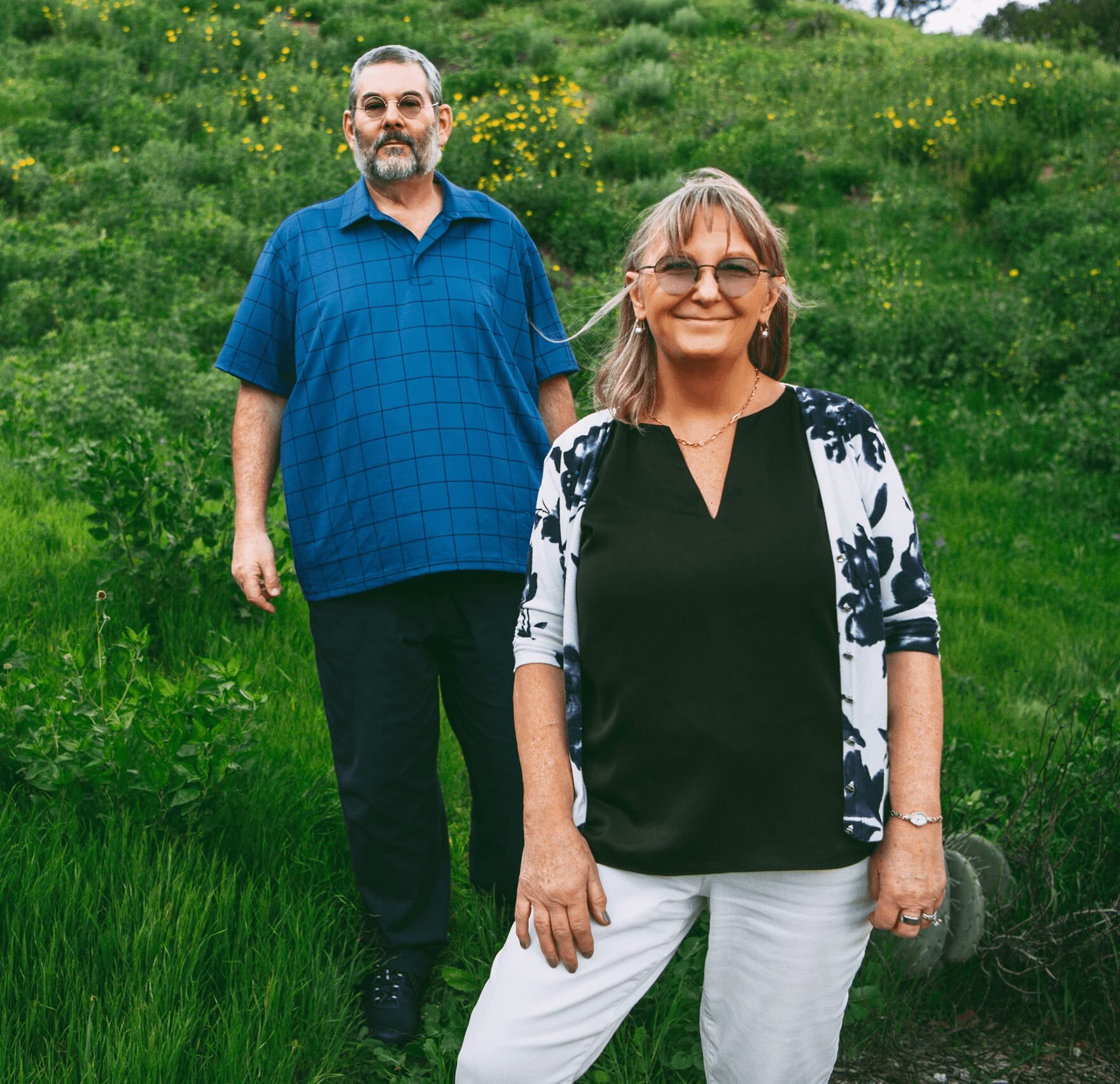A smiling woman in sunglasses and a black and white patterned top standing outdoors in a lush green field with yellow flowers, with a man in a blue checkered shirt and glasses standing behind her.