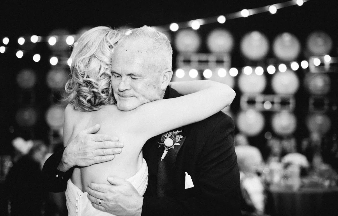 An older man in a suit hugging a woman. They are embracing closely, with the woman’s back to the camera and her arms around the man. The background shows blurred lights and a decorated setting, indicating a celebration or special event.