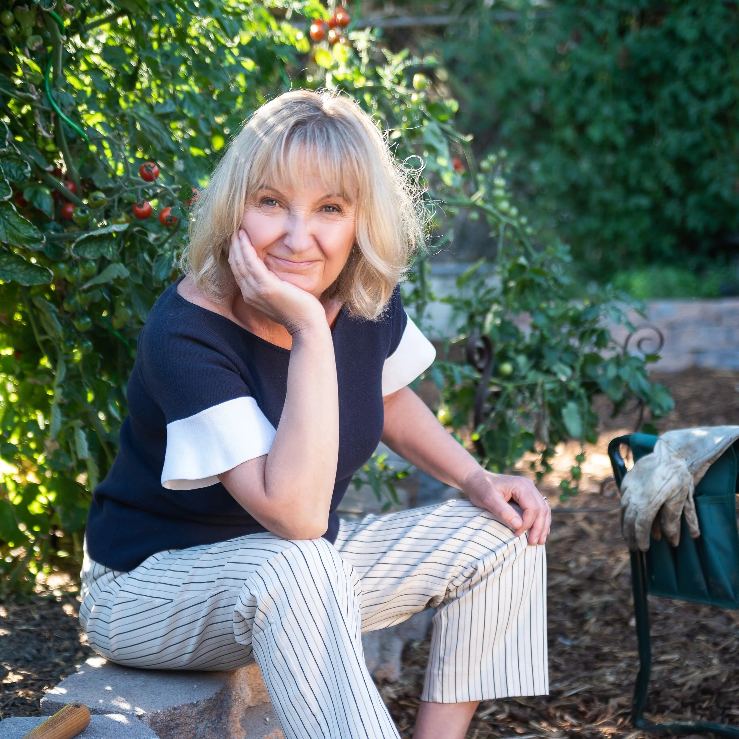 A woman sitting outdoors near lush green plants and tomato bushes, smiling and resting her chin on her hand.