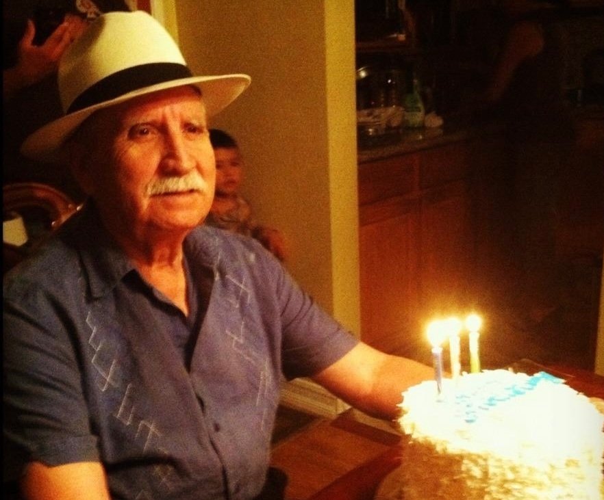 An elderly man with a white mustache wearing a white fedora hat and a blue short-sleeve shirt sitting at a table with a birthday cake that has lit candles.