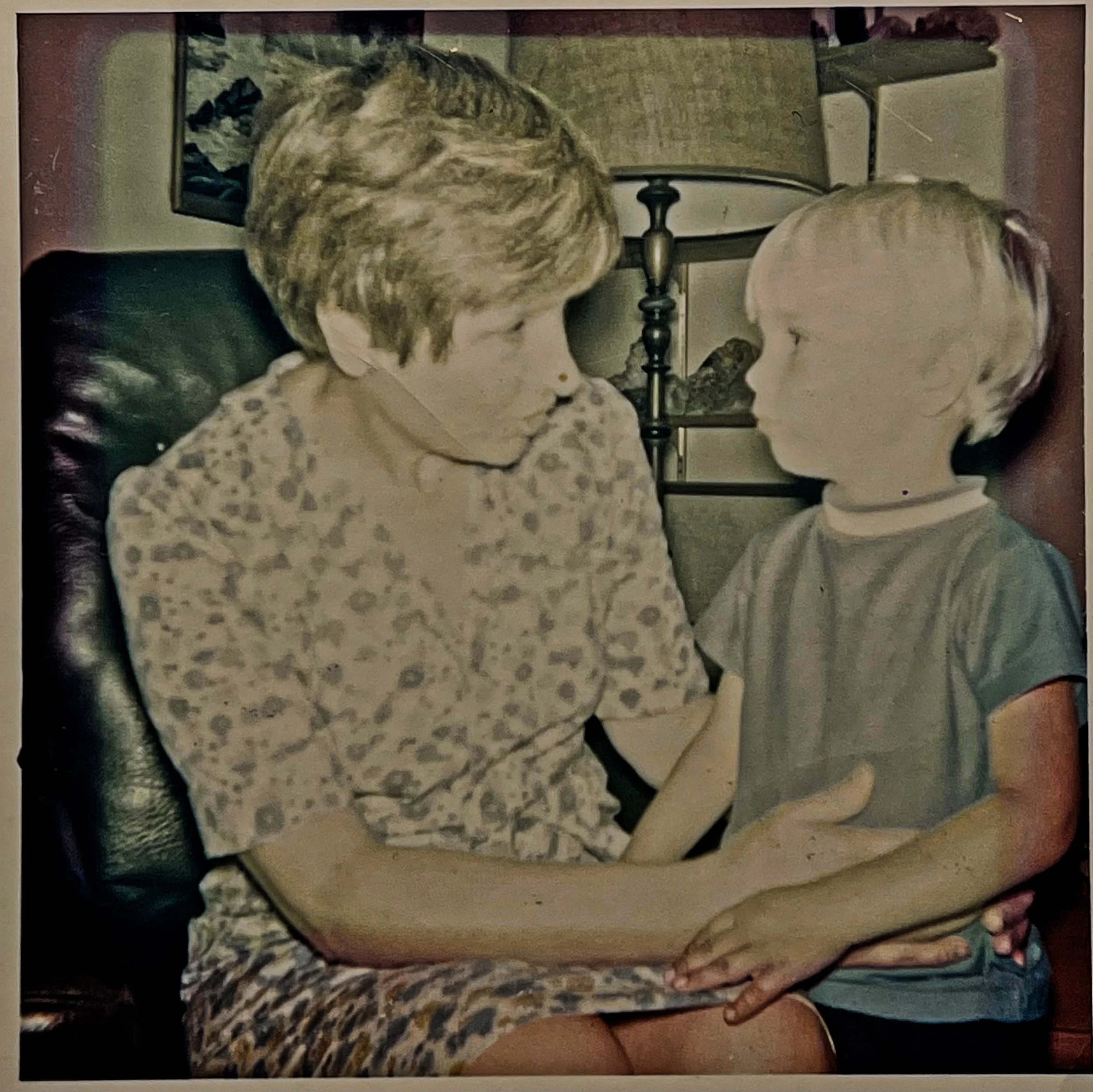 A woman and a young boy sit facing each other indoors. The woman has short hair and wears a patterned top, holding the boy's hand. The boy has light hair and wears a t-shirt, looking intently at the woman.