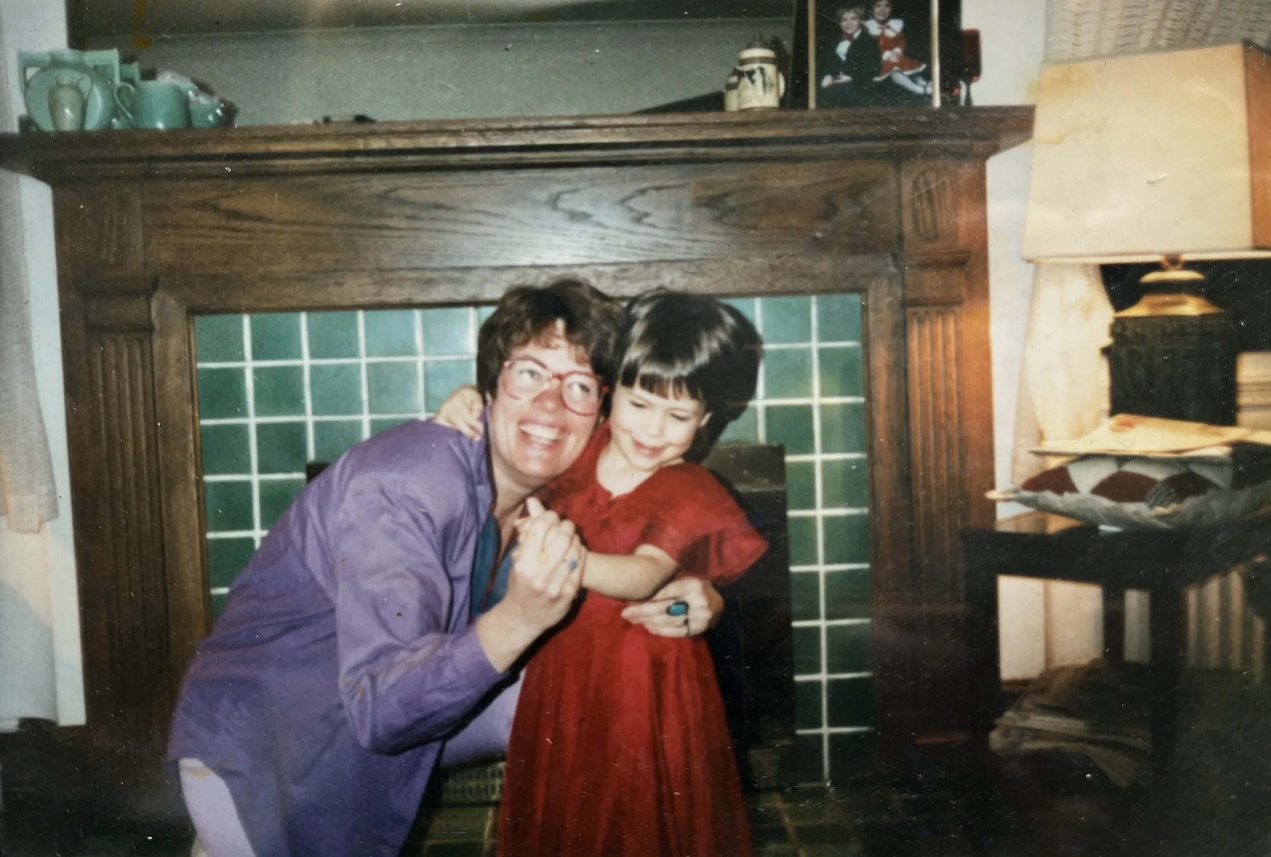 A woman and a young girl smiling and hugging near a fireplace in a cozy living room.