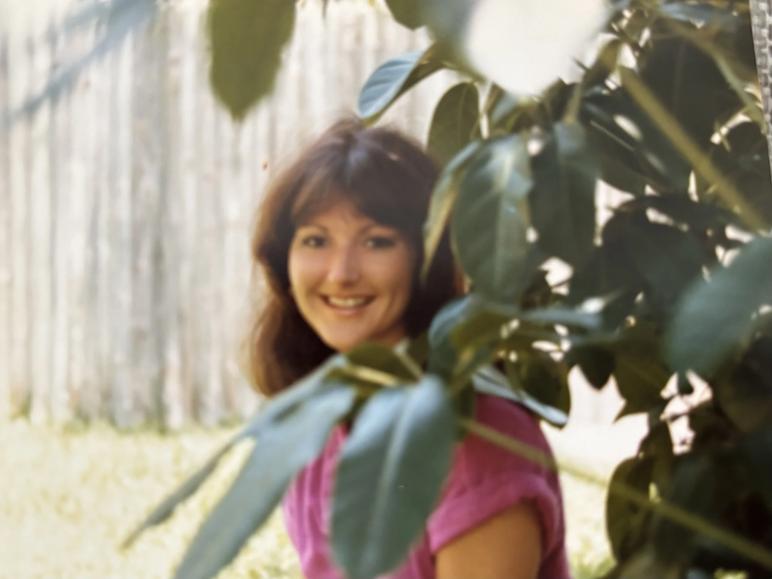 A woman with shoulder-length brown hair smiling outdoors, partially obscured by green leaves in the foreground, with a wooden fence in the background.