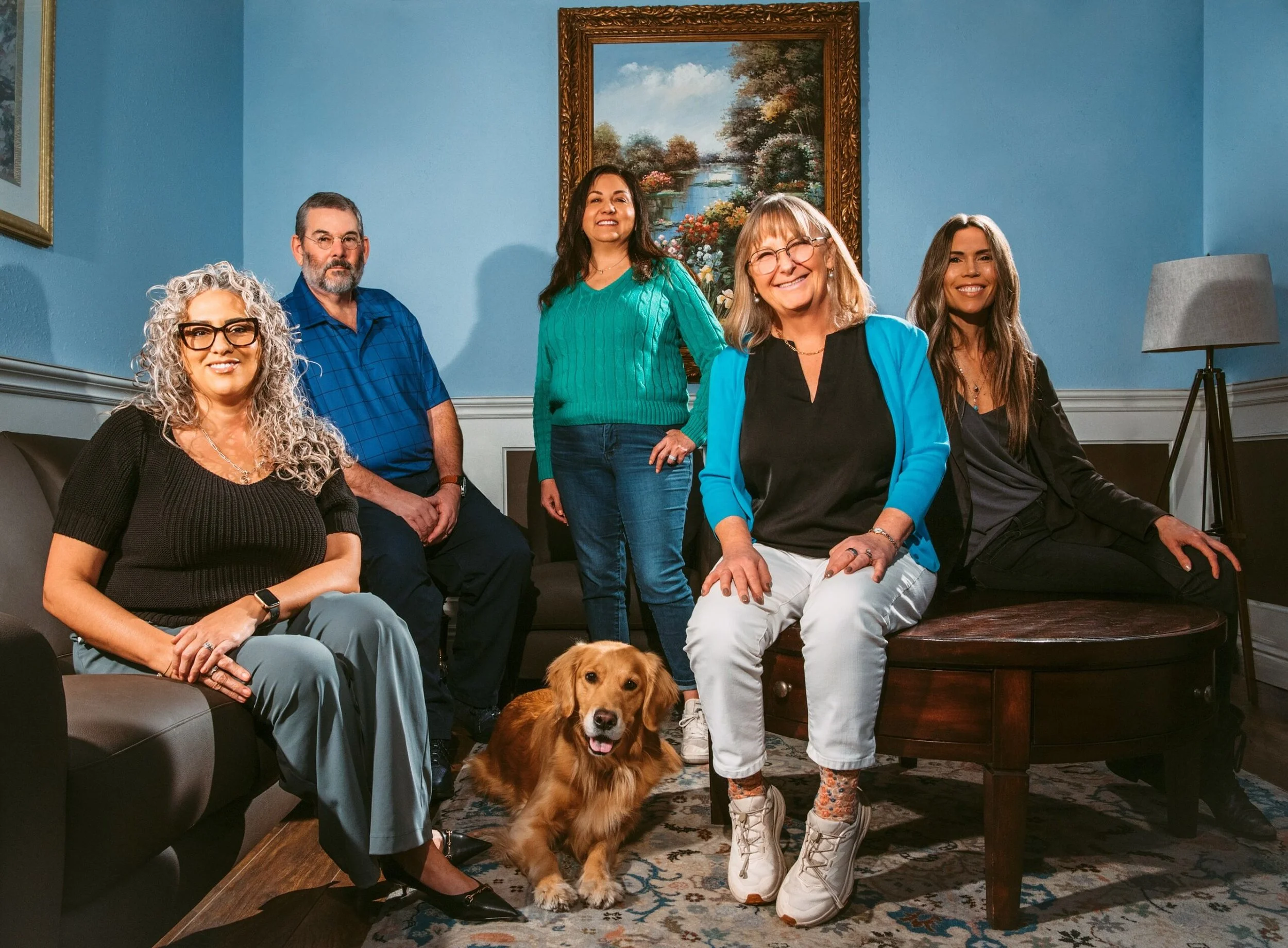 A group of six adults and one dog in a living room. The photo shows three women and one man standing and sitting, with a woman and a man sitting on a couch. The dog, a Cocker Spaniel, is sitting on the floor in front of the group. The background features a painting of a landscape with water and trees, blue walls, a lamp, and a patterned rug.
