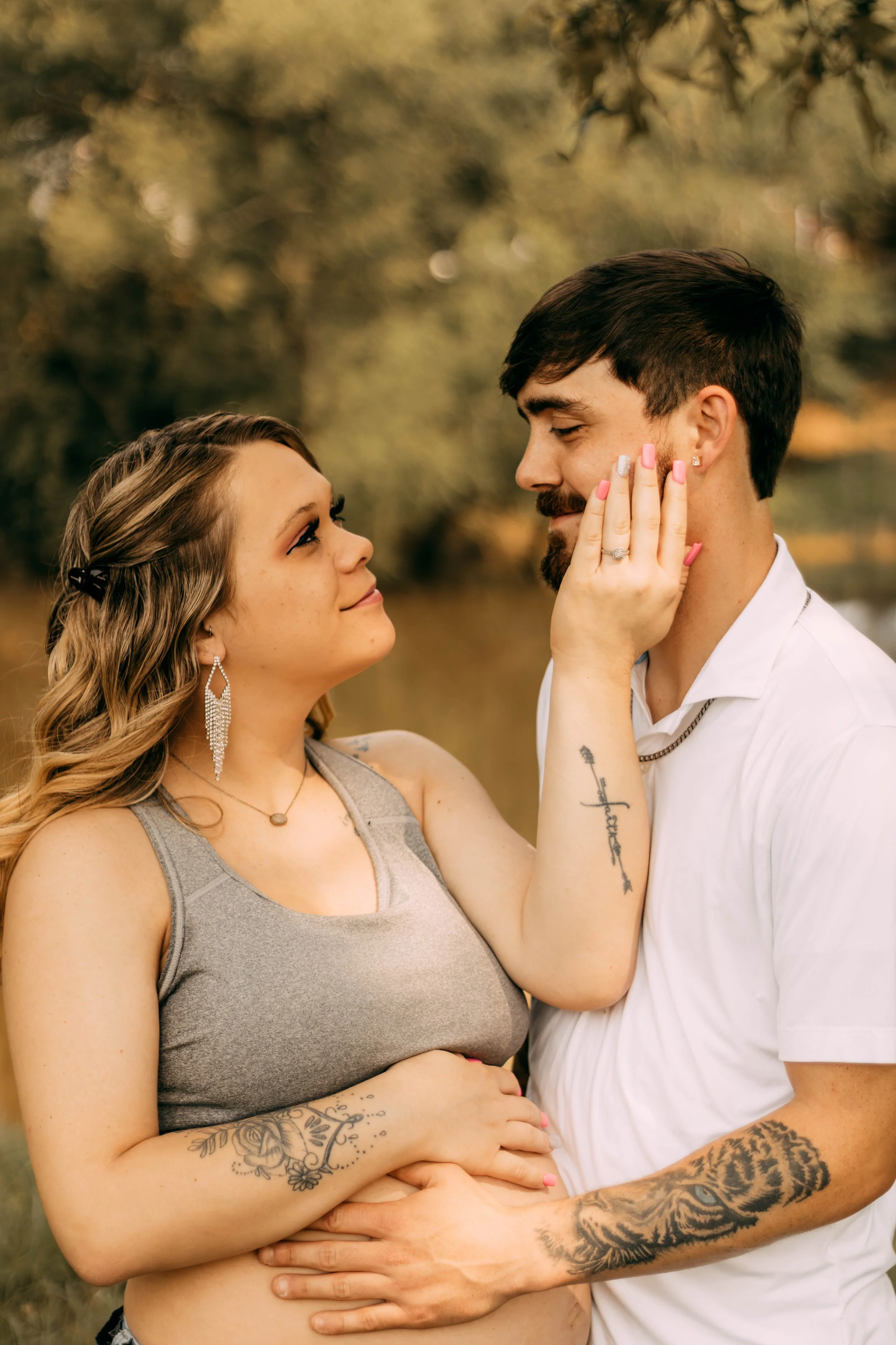 A couple, woman with long wavy hair and man with short dark hair, sharing an intimate moment outdoors in a park. The woman touches the man's face affectionately, and they look into each other's eyes. Both have tattoos and wear casual clothes.