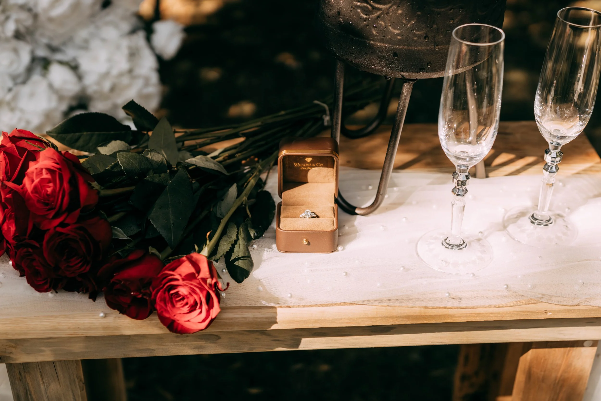 A wooden table with a bouquet of red roses, an open ring box with a diamond ring, two empty champagne flutes, and a grill in the background.