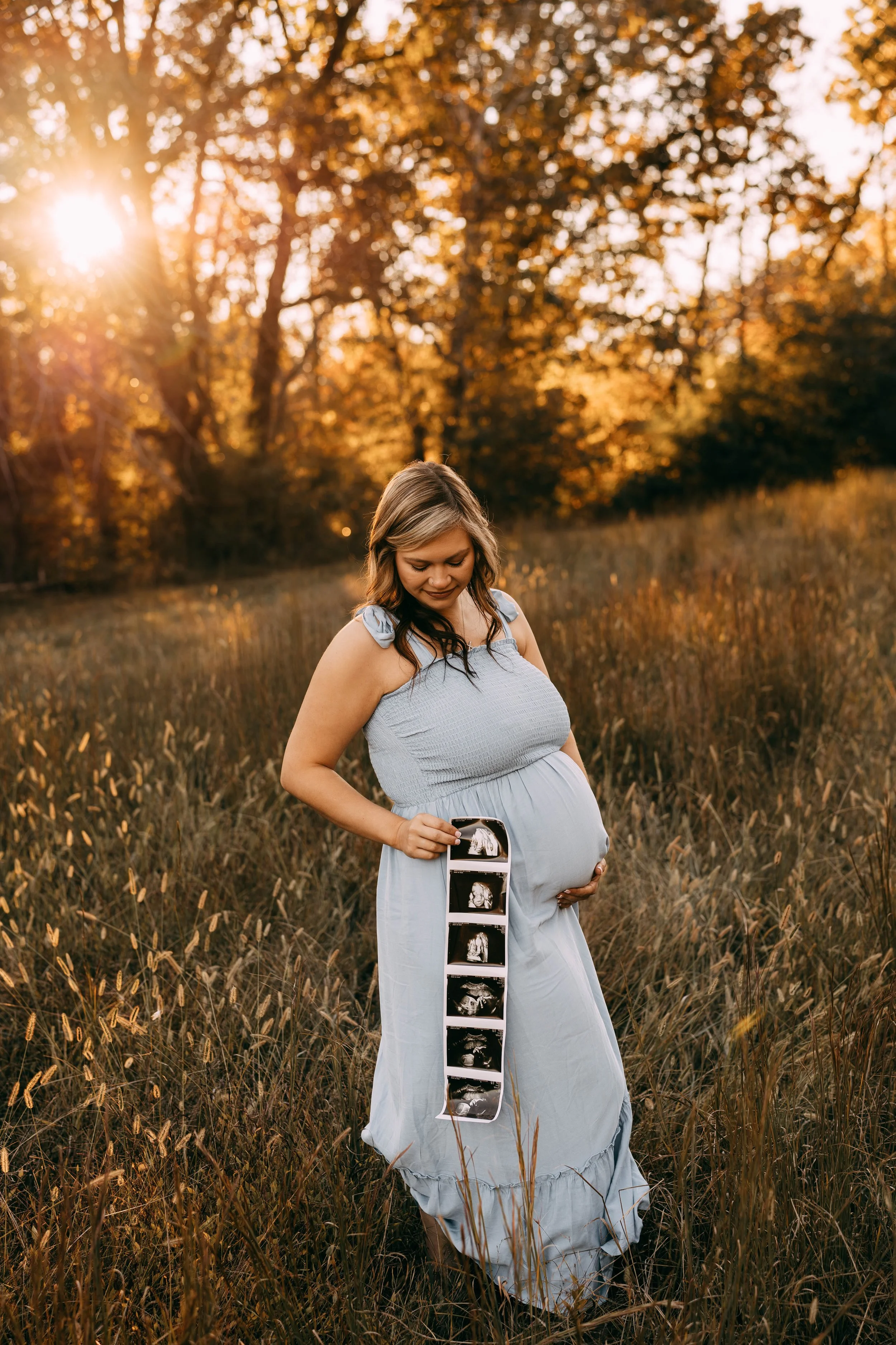 Pregnant woman in a meadow holding ultrasound scans, with the setting sun and trees in the background.