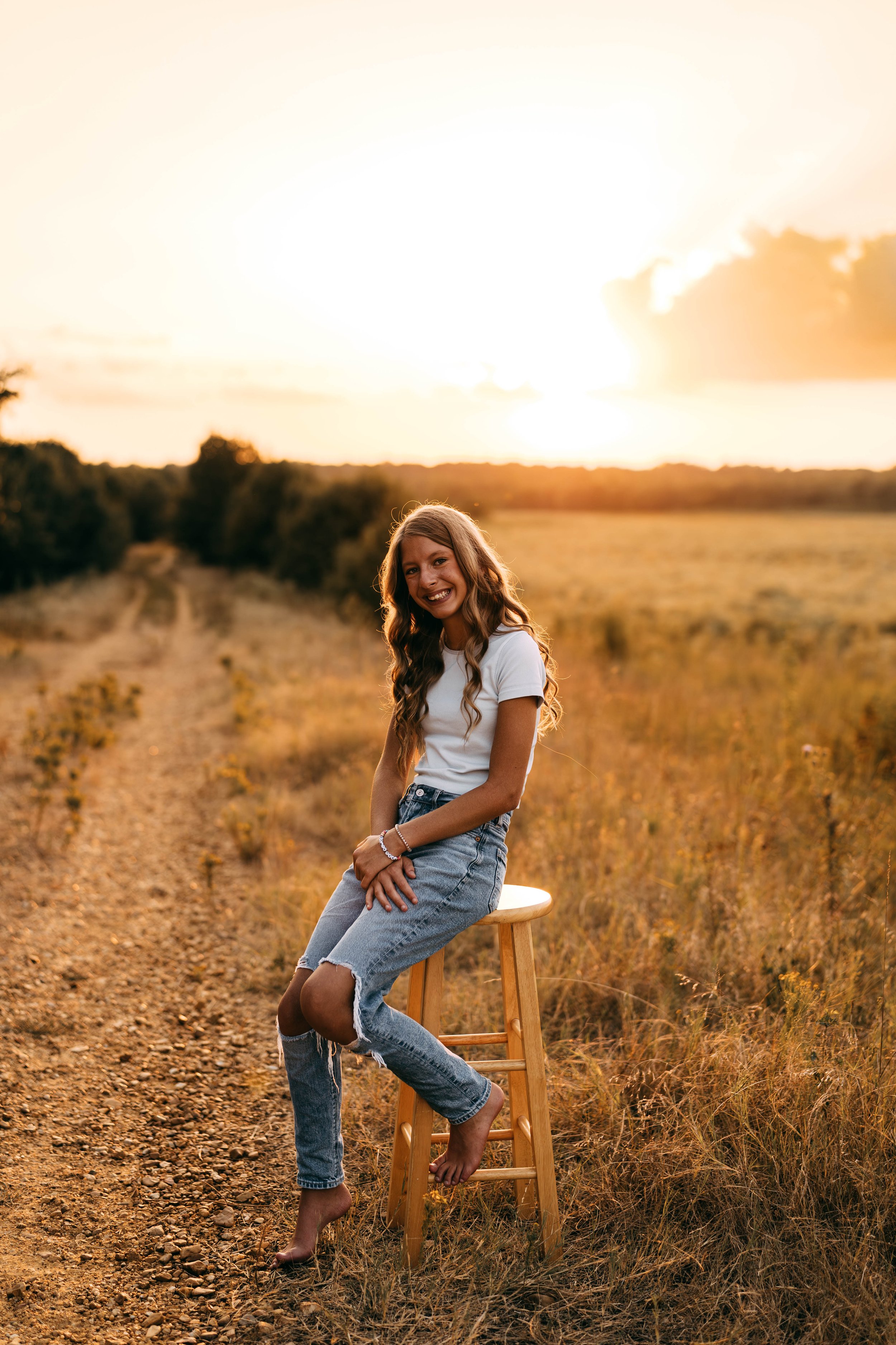 A young woman sitting on a wooden stool in a rural field at sunset, smiling and wearing a white t-shirt and ripped jeans.