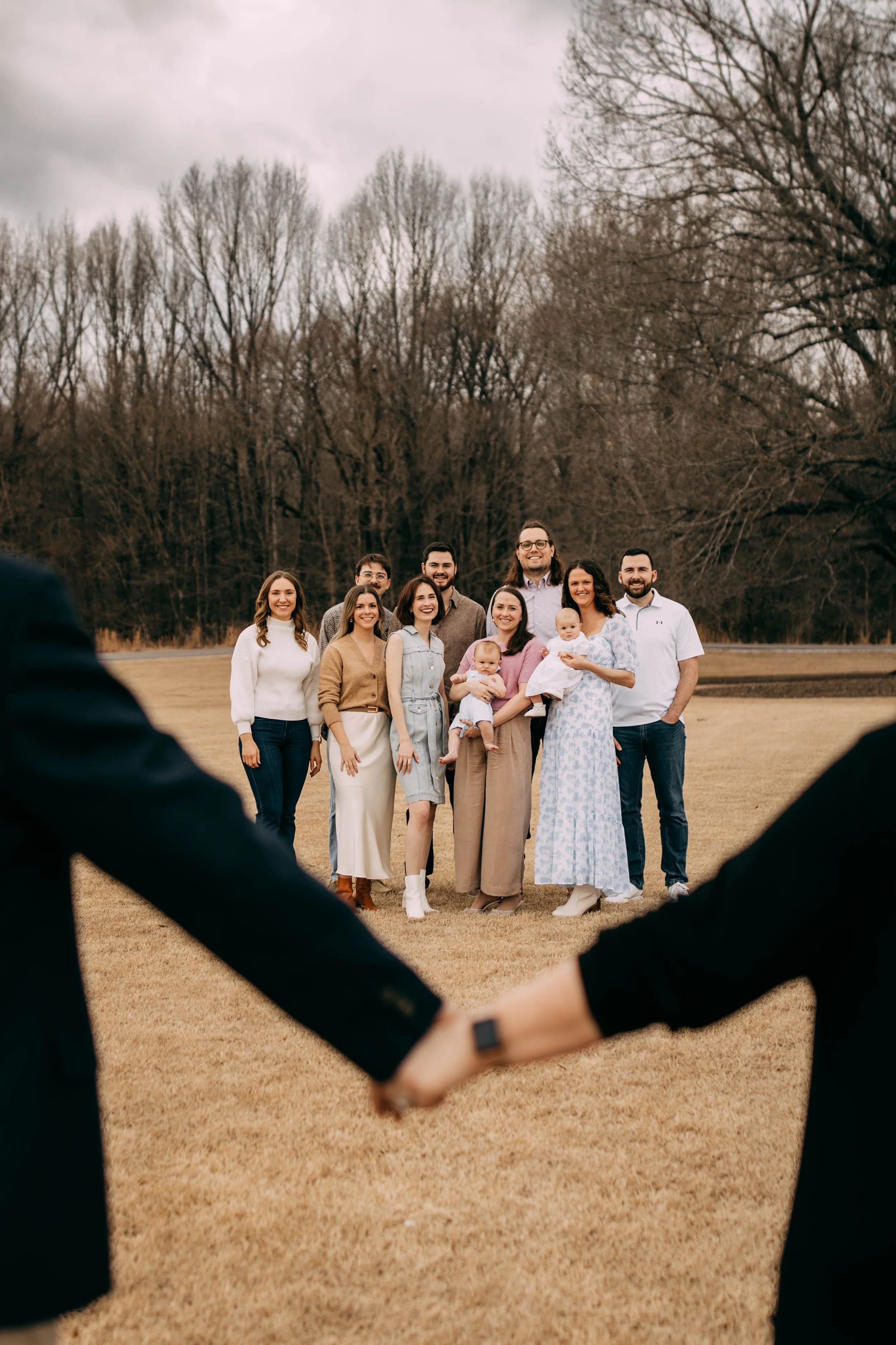 Group of friends and family standing on a grassy field, posing for a photo during daytime, with two people holding hands in the foreground.